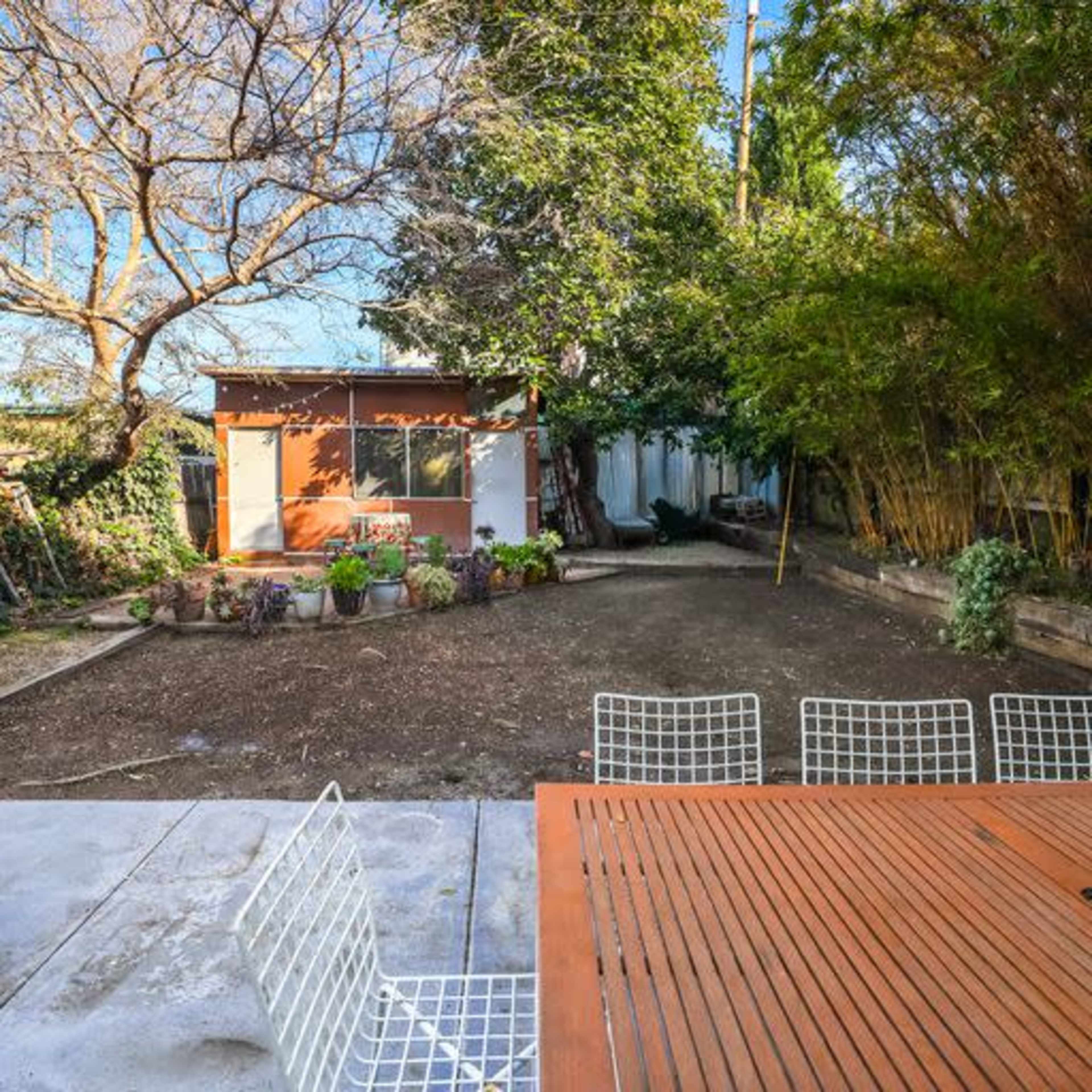 A backyard features a wooden table with white chairs in the foreground, a small garden area, and a shed surrounded by trees in the background.