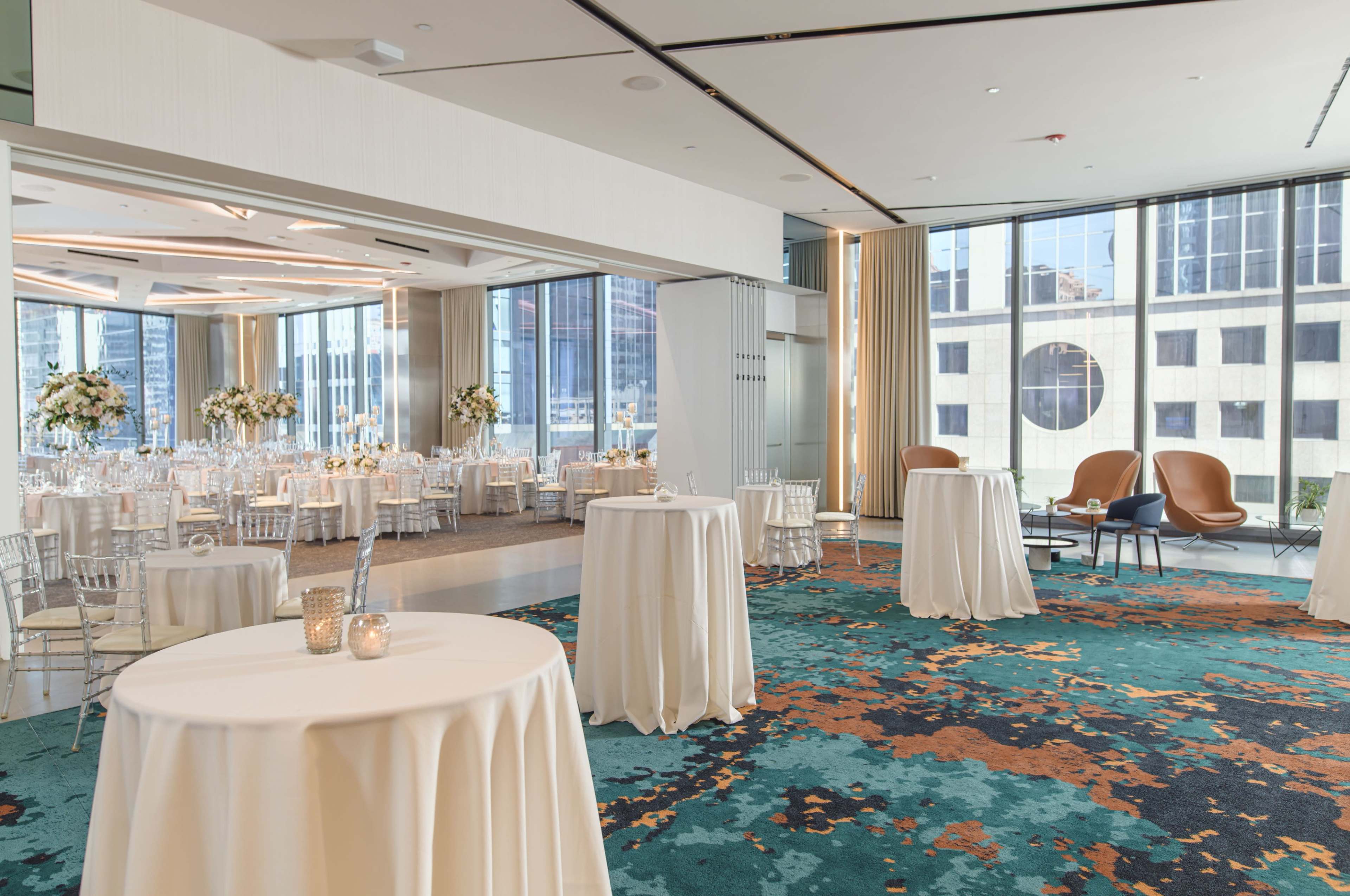The image shows a spacious event venue with round tables covered in white tablecloths, elegant seating, and large windows showcasing a city view.