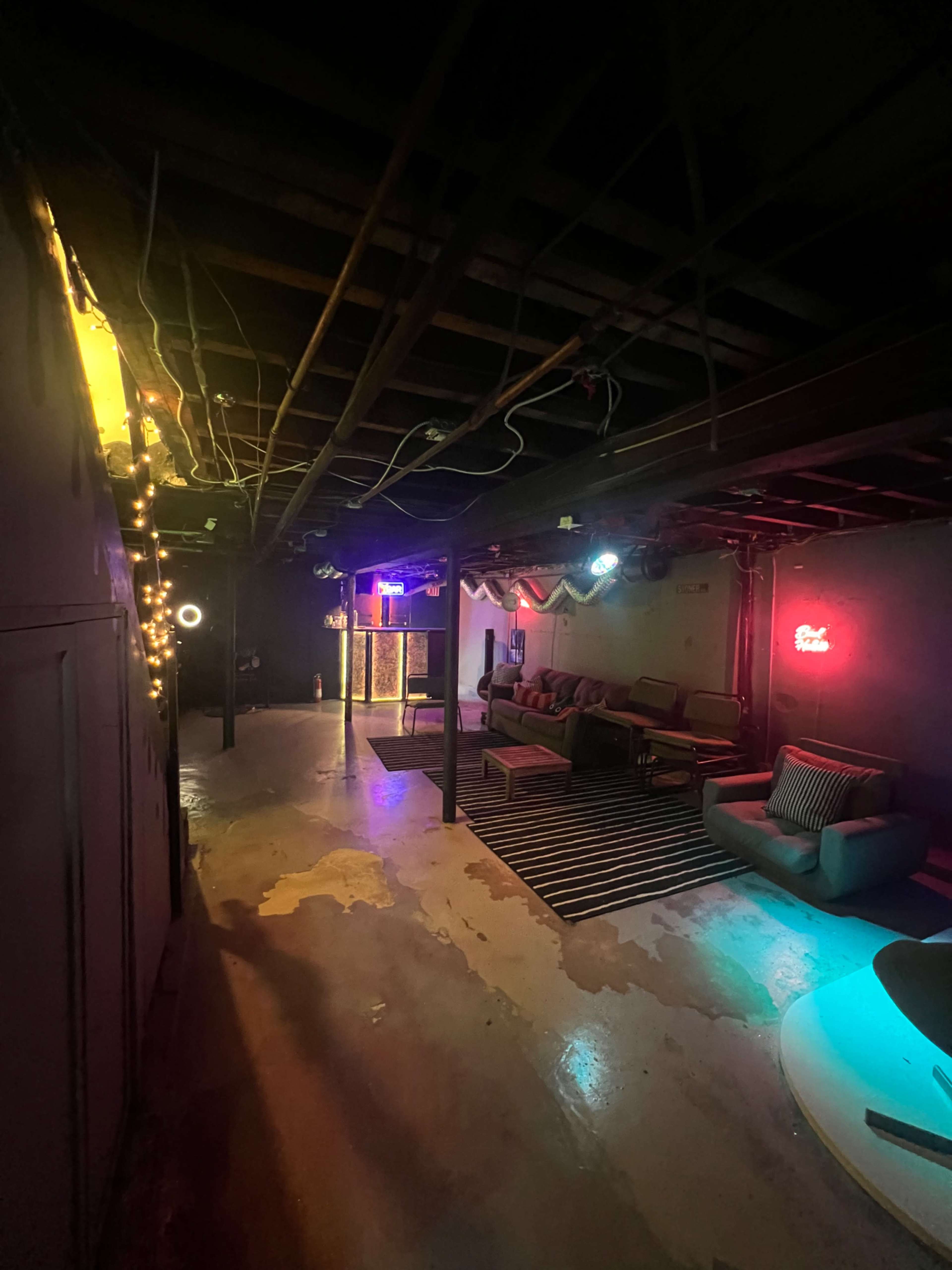 A dimly lit basement features lounge furniture, neon lights, and decorative string lights along the walls.