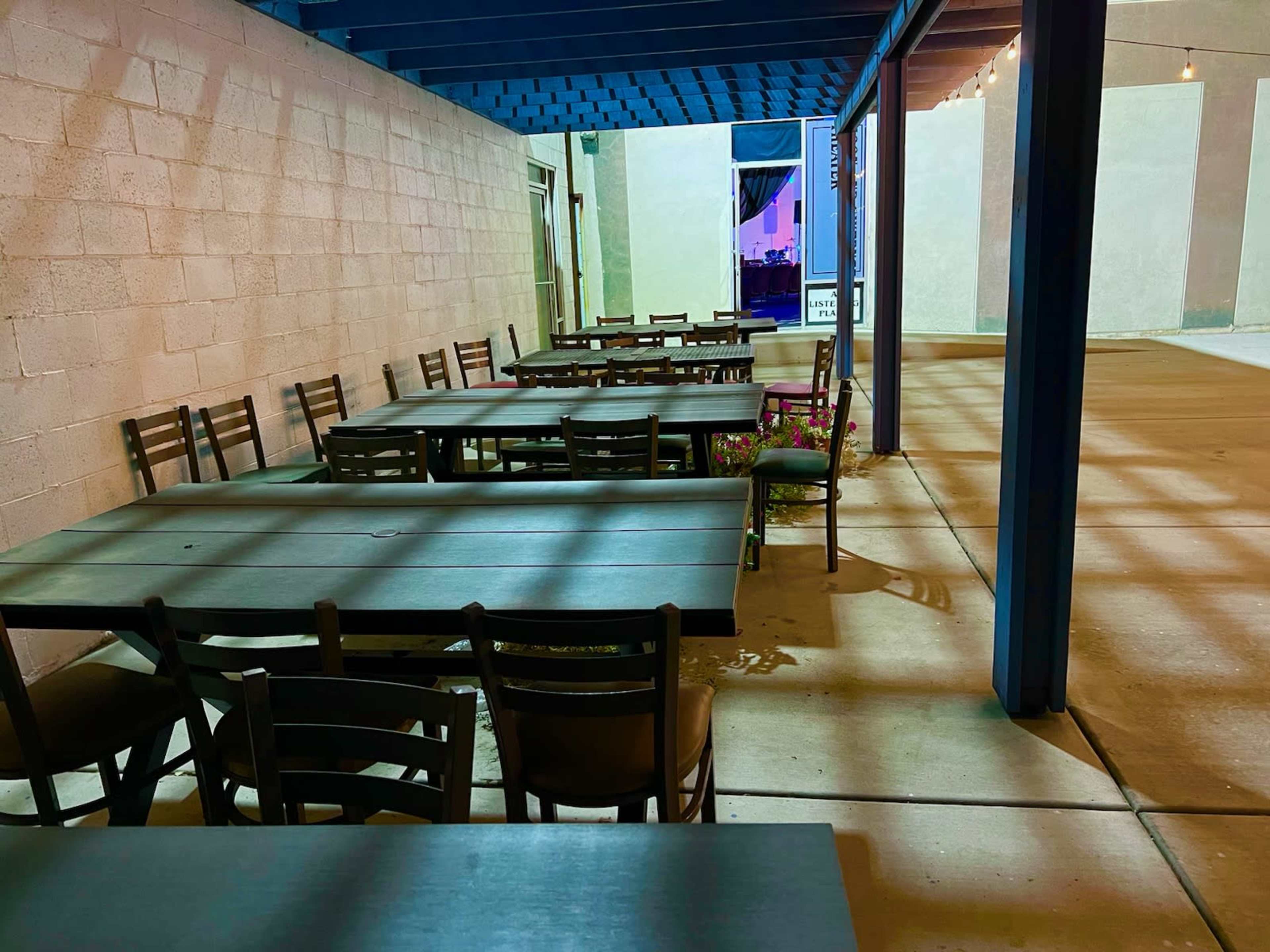 An outdoor dining area features several empty tables and chairs under a blue overhead structure, with light illuminating the surrounding space.