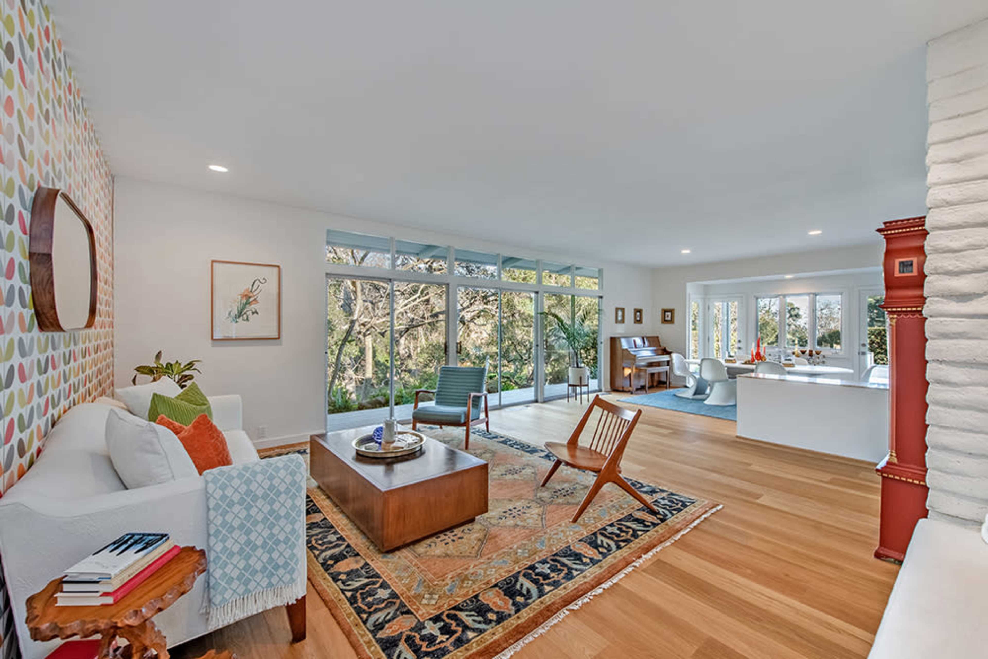 The image shows a modern living room with a patterned accent wall, a wooden coffee table, and large windows overlooking a wooded area.