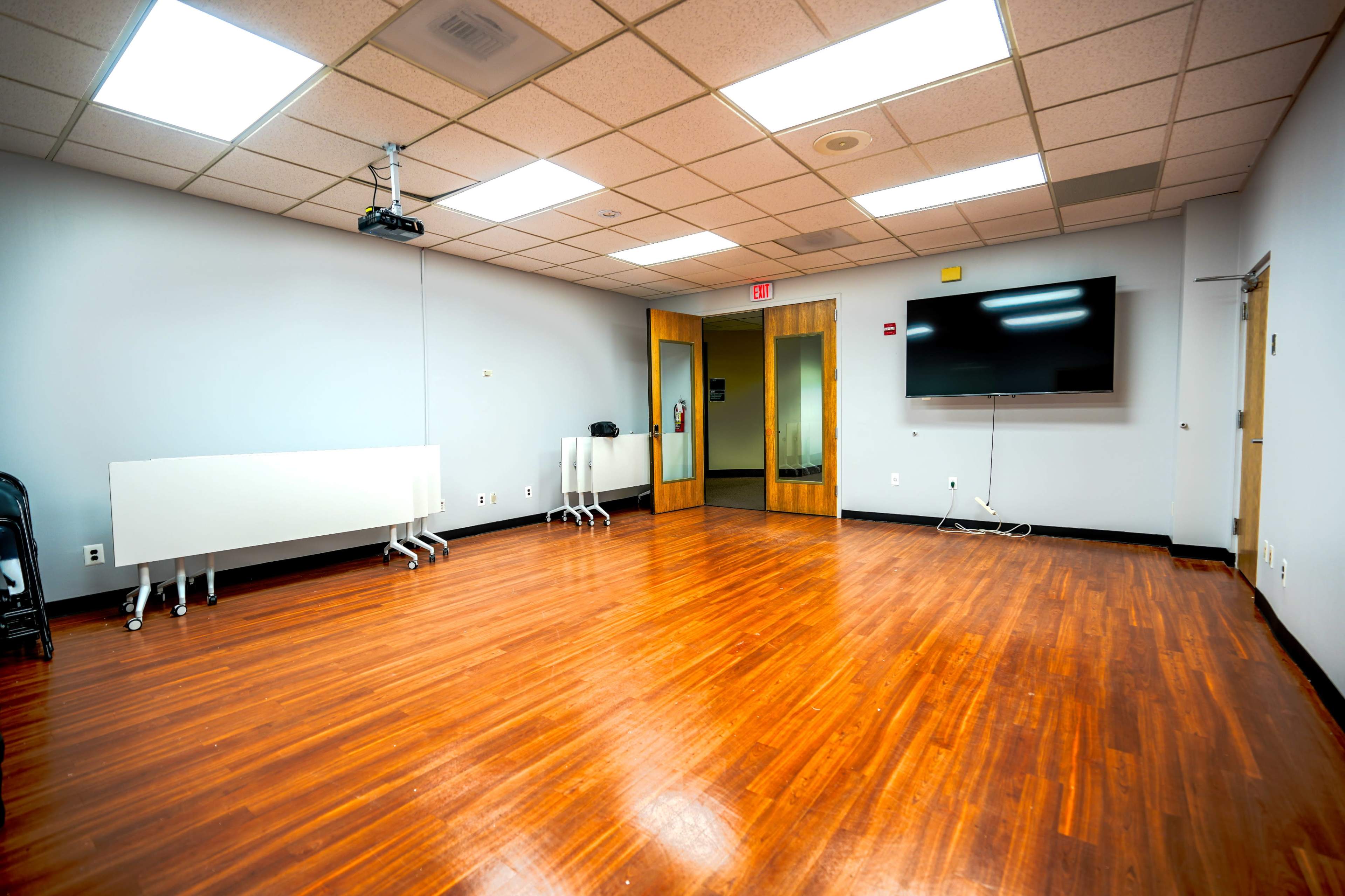The image shows a vacant room with wooden flooring, featuring a wall-mounted TV, a projector, and two folding tables stored against the walls.