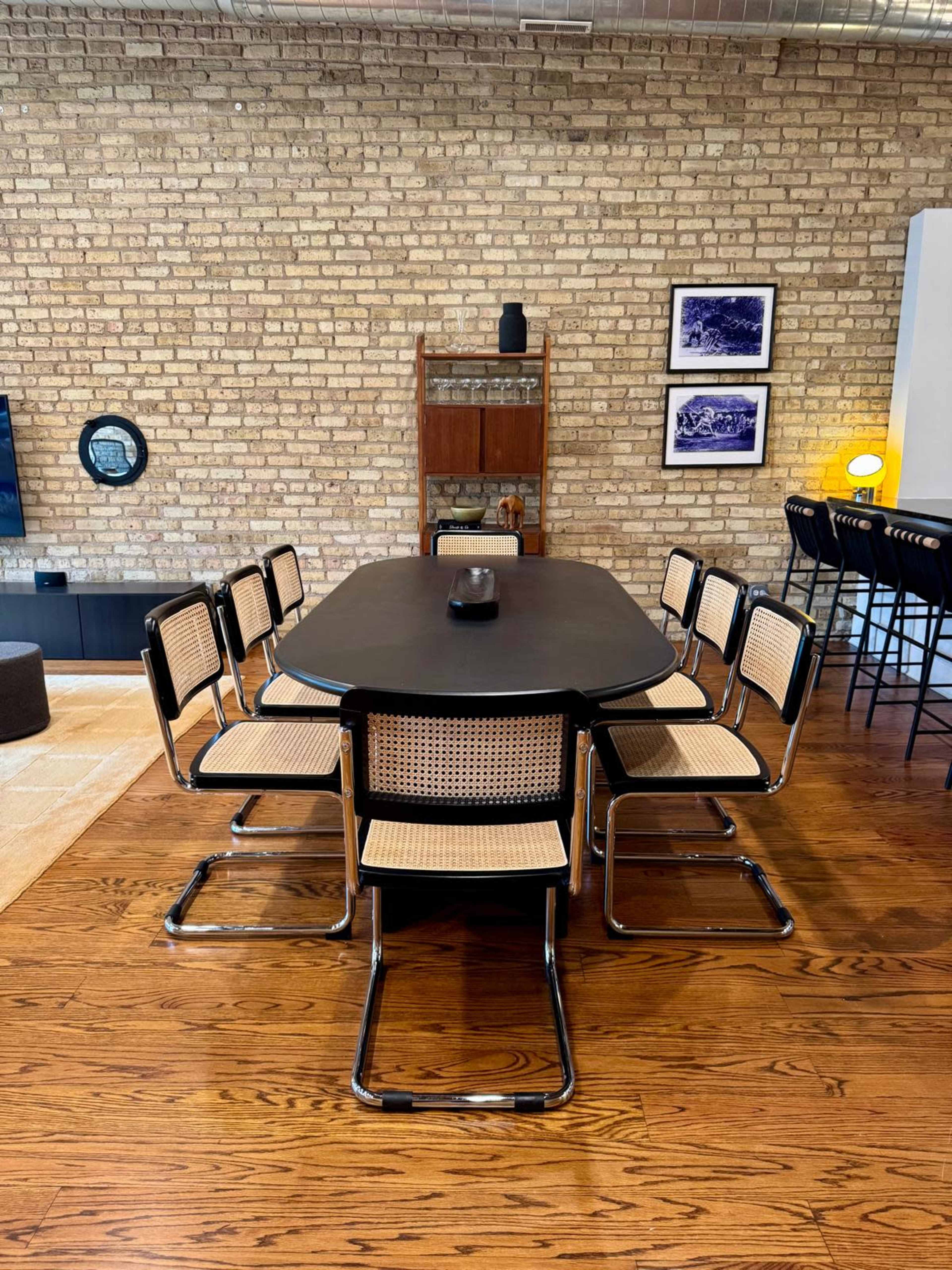 A rectangular black dining table surrounded by six cane-backed chairs is set against a brick wall in a modern interior.