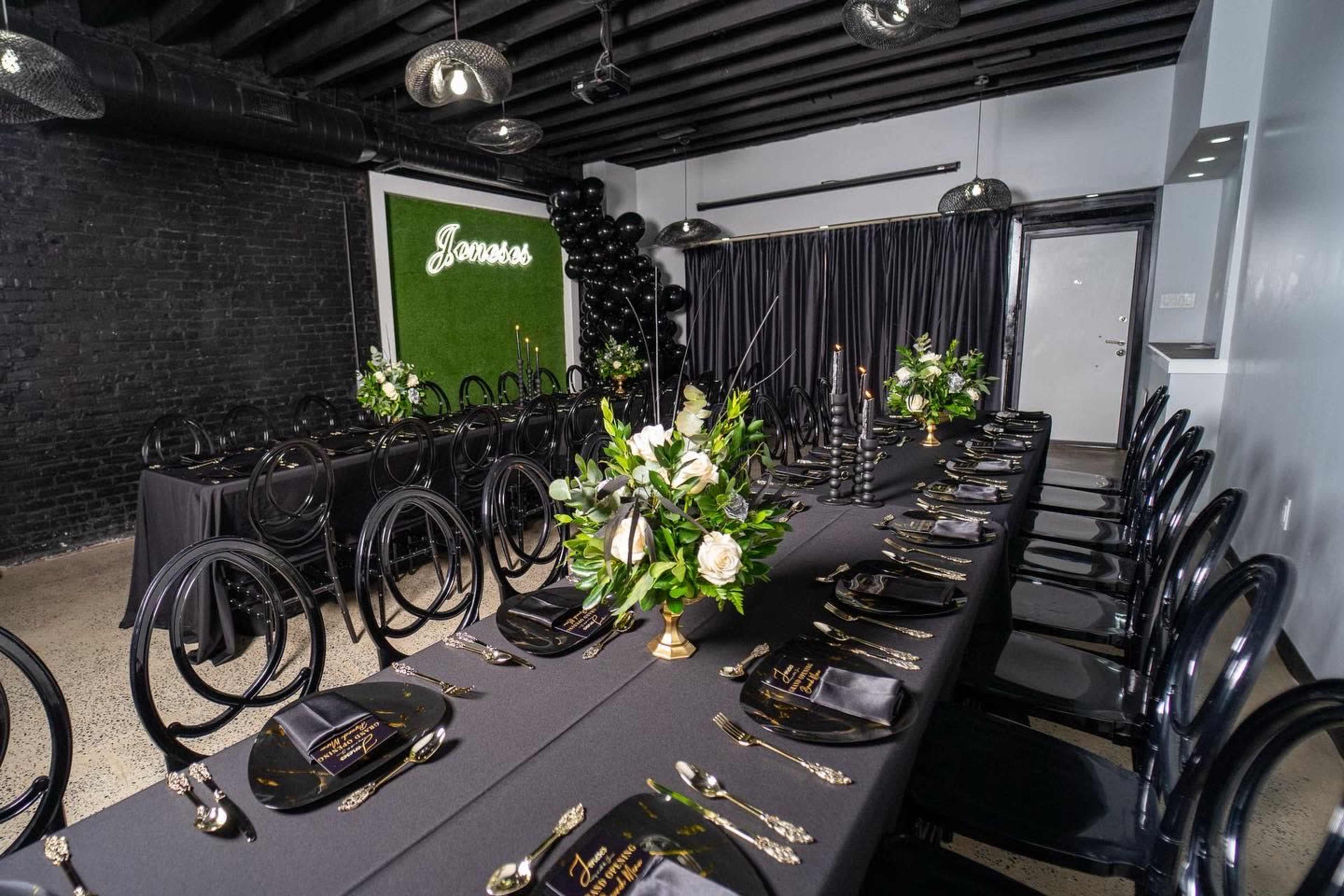 A long, elegantly set dining table with black tablecloths, black chairs, and floral centerpieces is arranged in a decorated room featuring black walls and greenery accents.