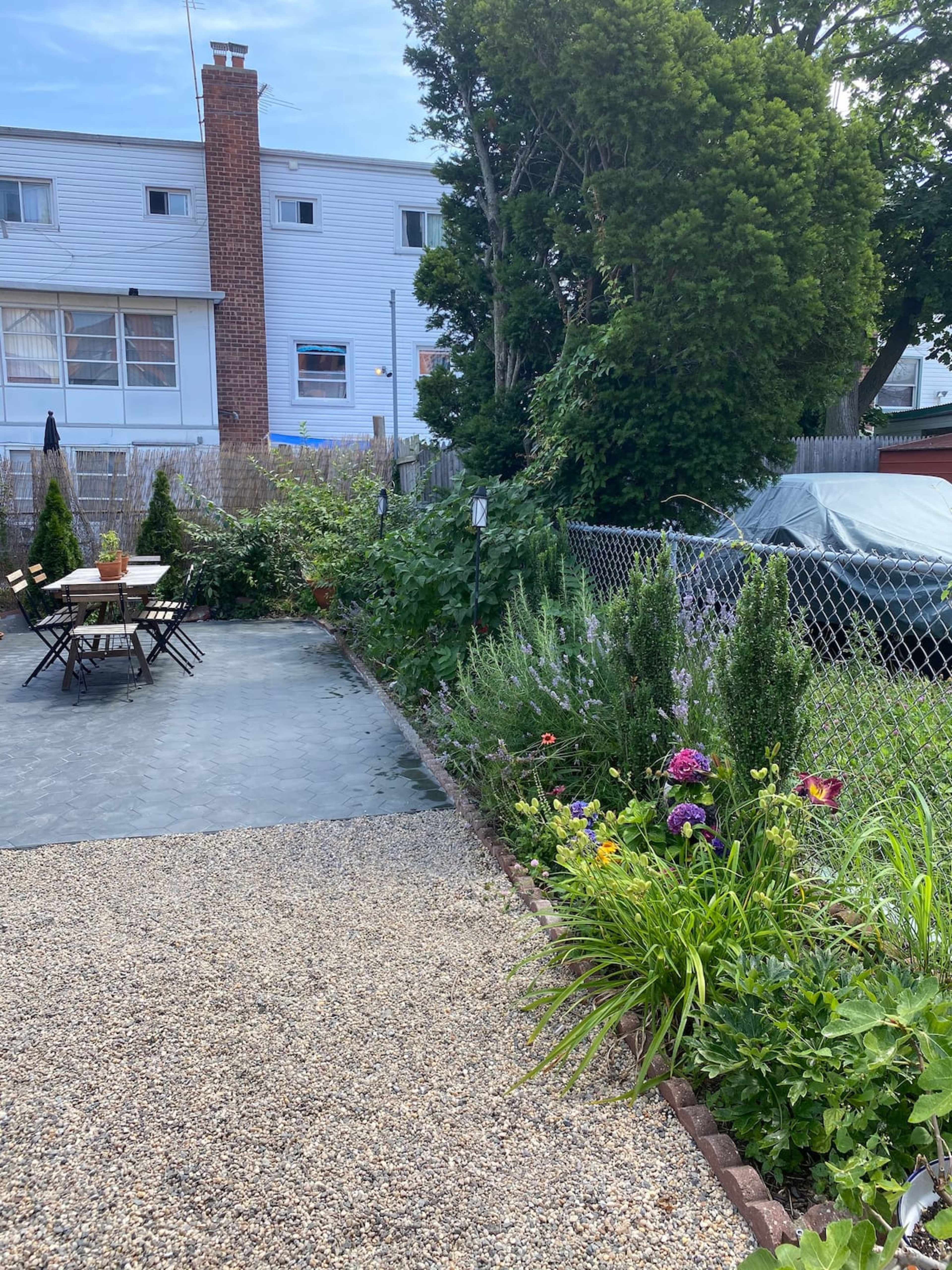 A backyard with a stone pathway leading to a table and chairs, surrounded by greenery and flowers, with a car partially covered in a tarp nearby.