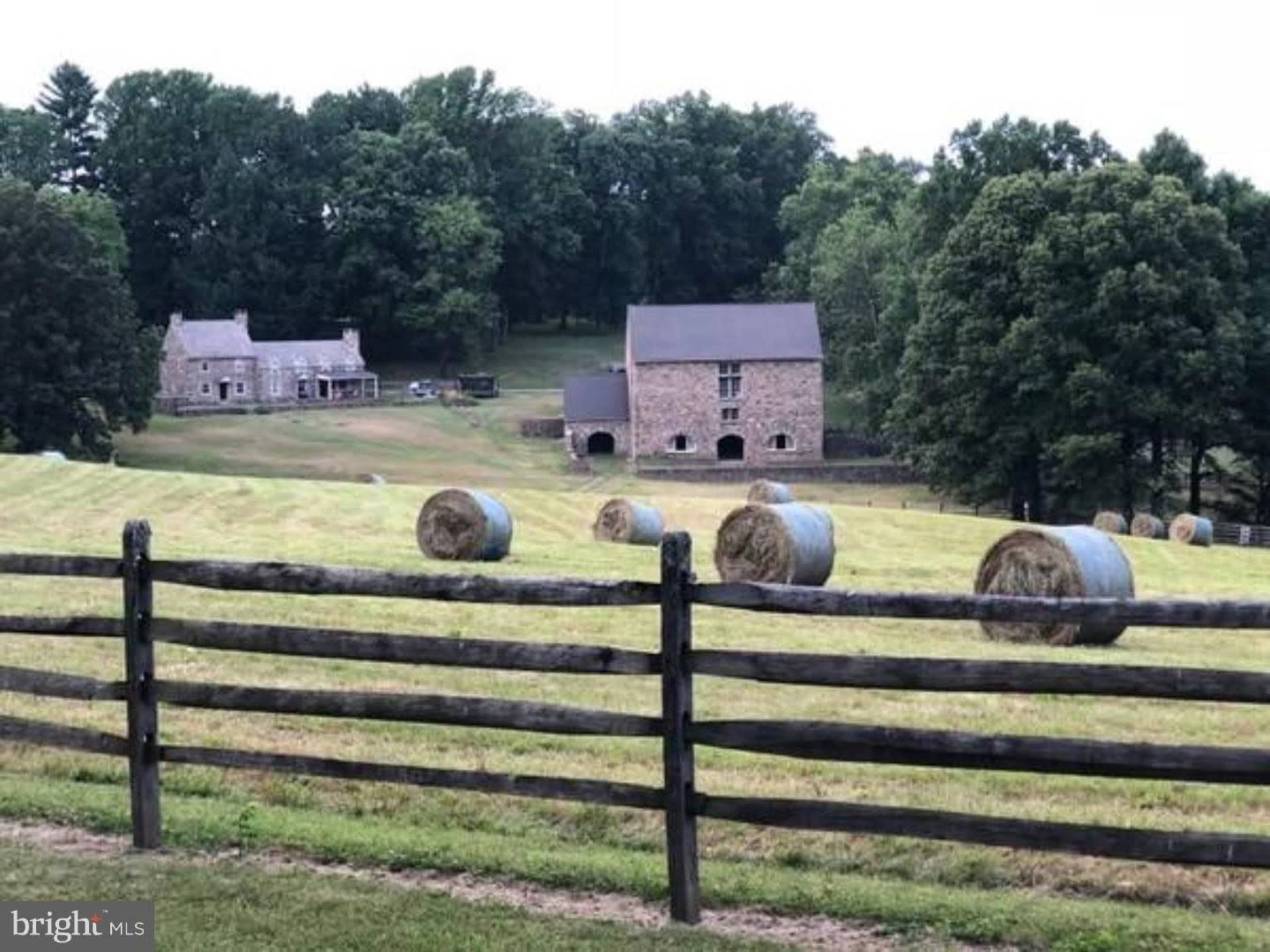 A stone farmhouse and barn are set against a backdrop of trees, with round hay bales scattered across a grassy field and a wooden fence in the foreground.