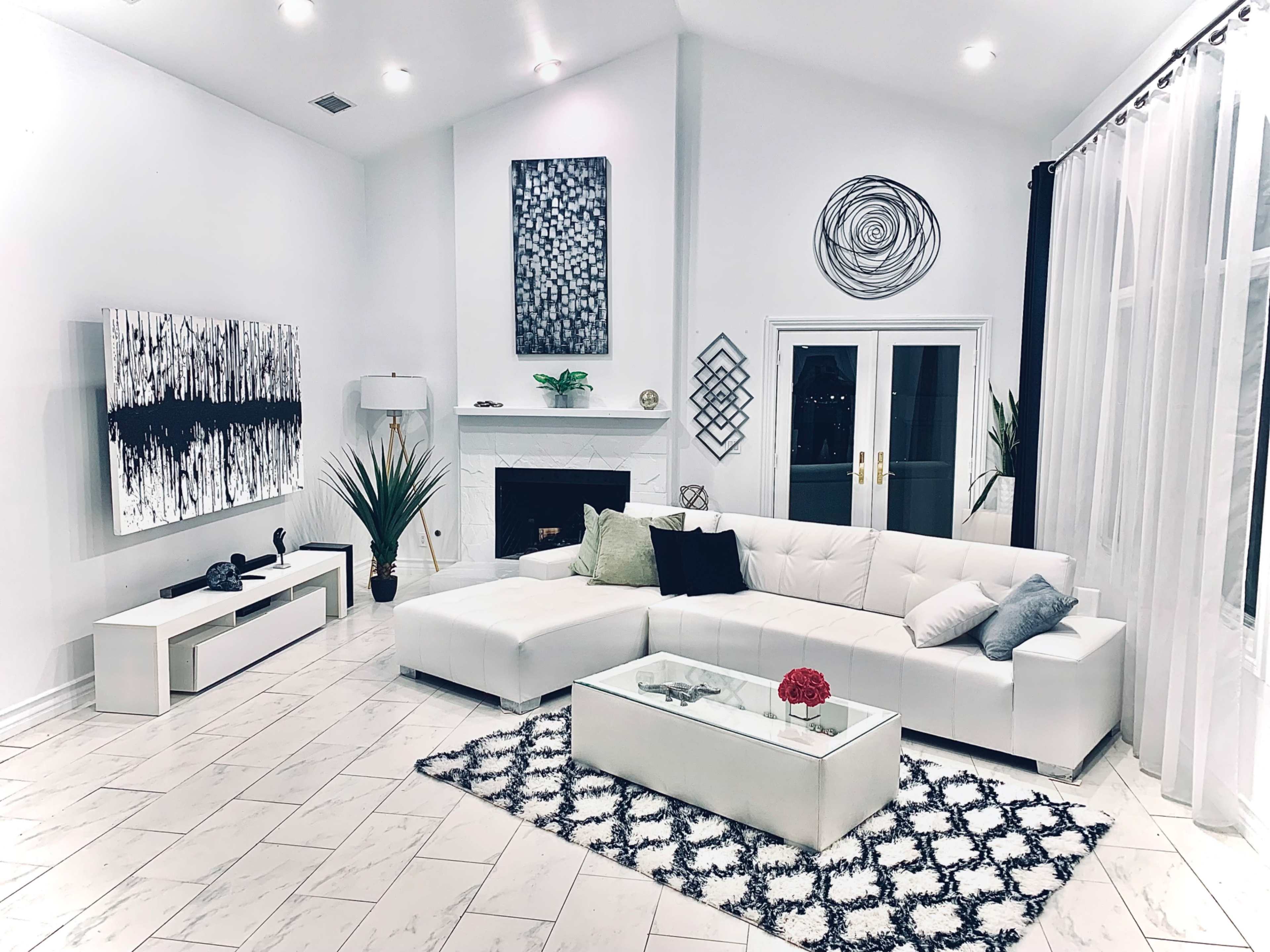 The image shows a modern living room featuring a white sectional sofa, a glass coffee table with a decorative item, and a fireplace, with abstract artwork on the walls and patterned rugs on the floor.
