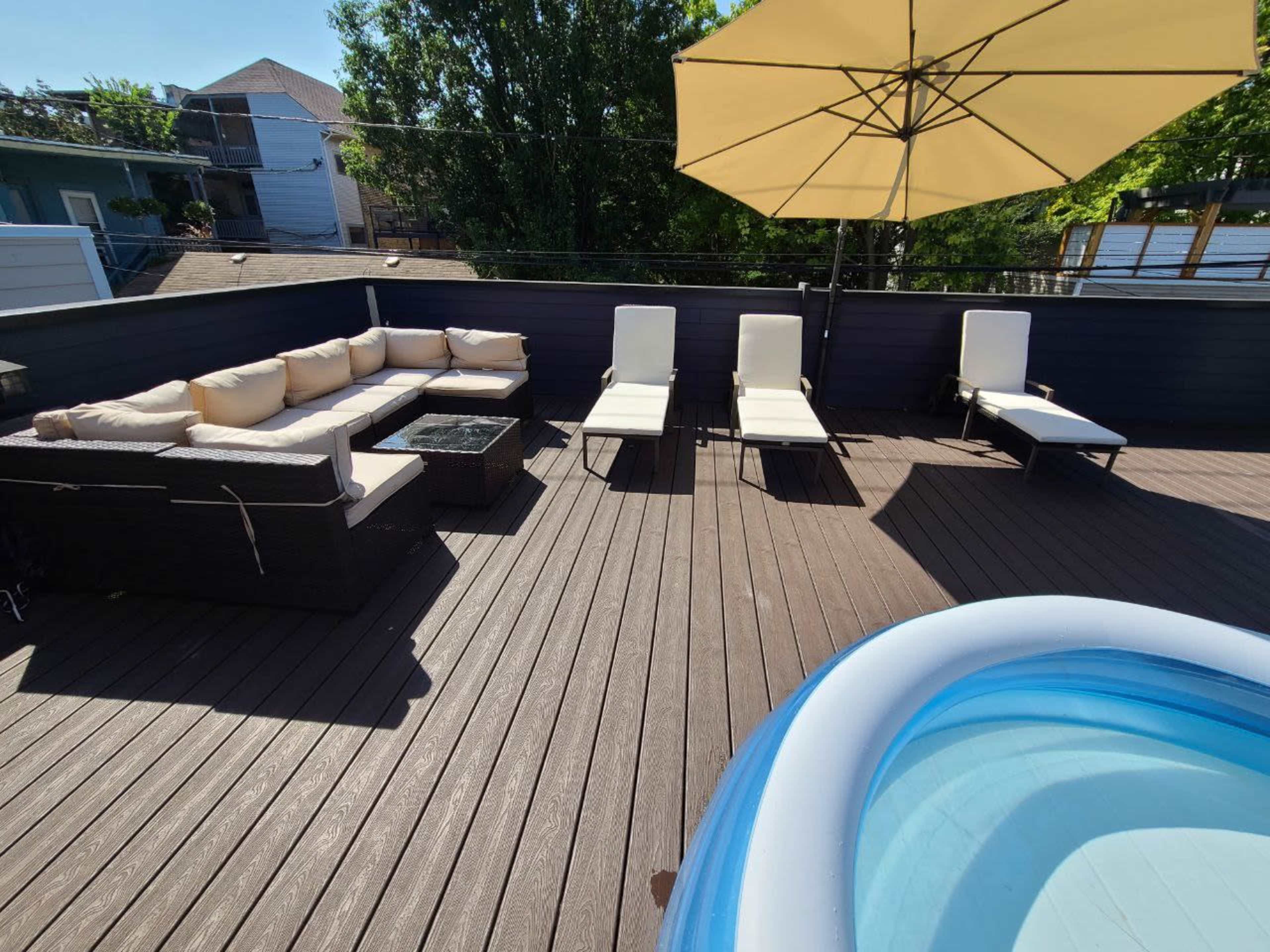 A rooftop terrace furnished with a sectional sofa, several lounge chairs, a small coffee table, and an inflatable hot tub, all under a large umbrella.