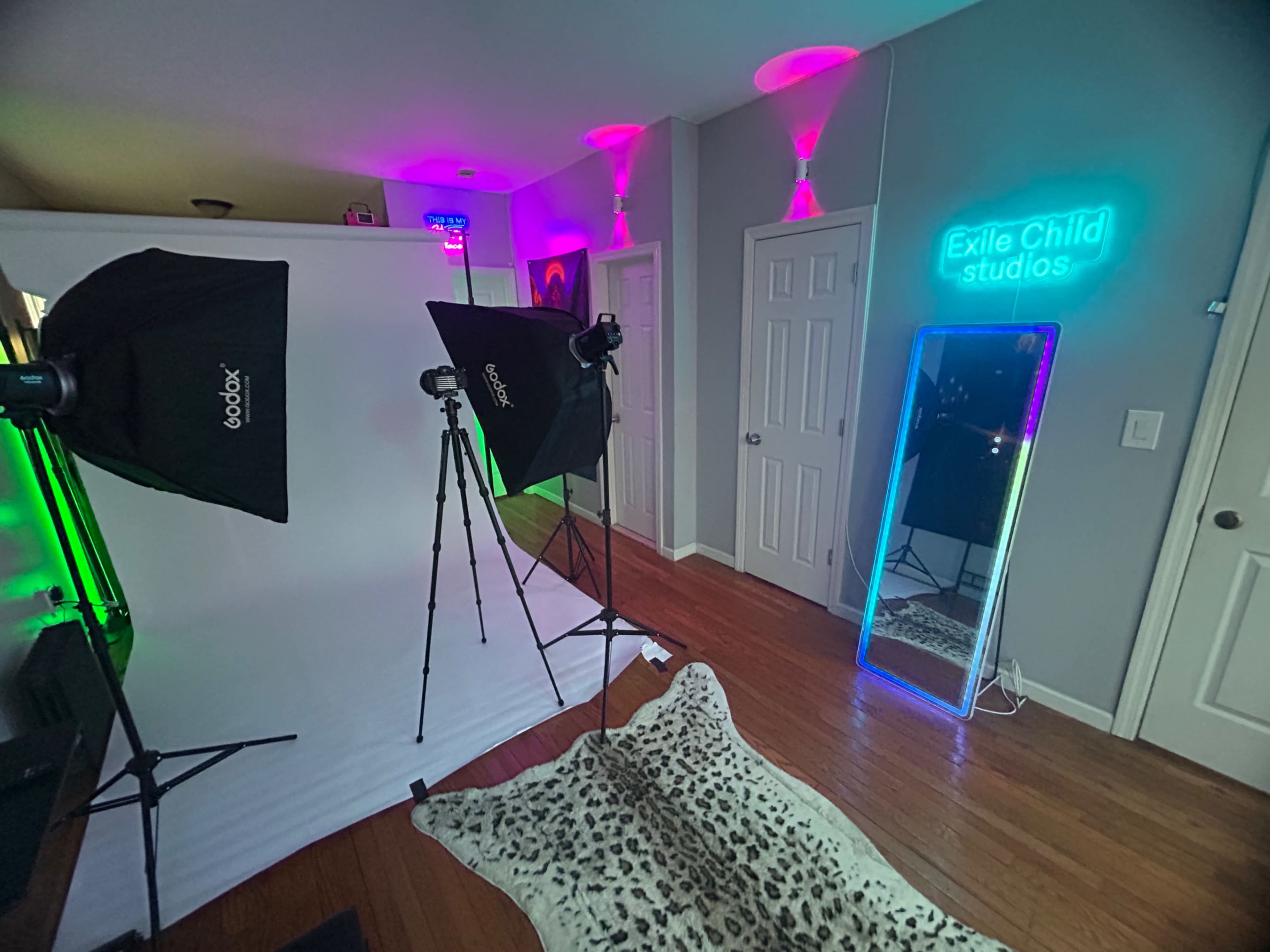 The image shows a photography studio setup featuring two softbox lights, a white backdrop, a neon studio sign, and a mirror in a room with wooden flooring.