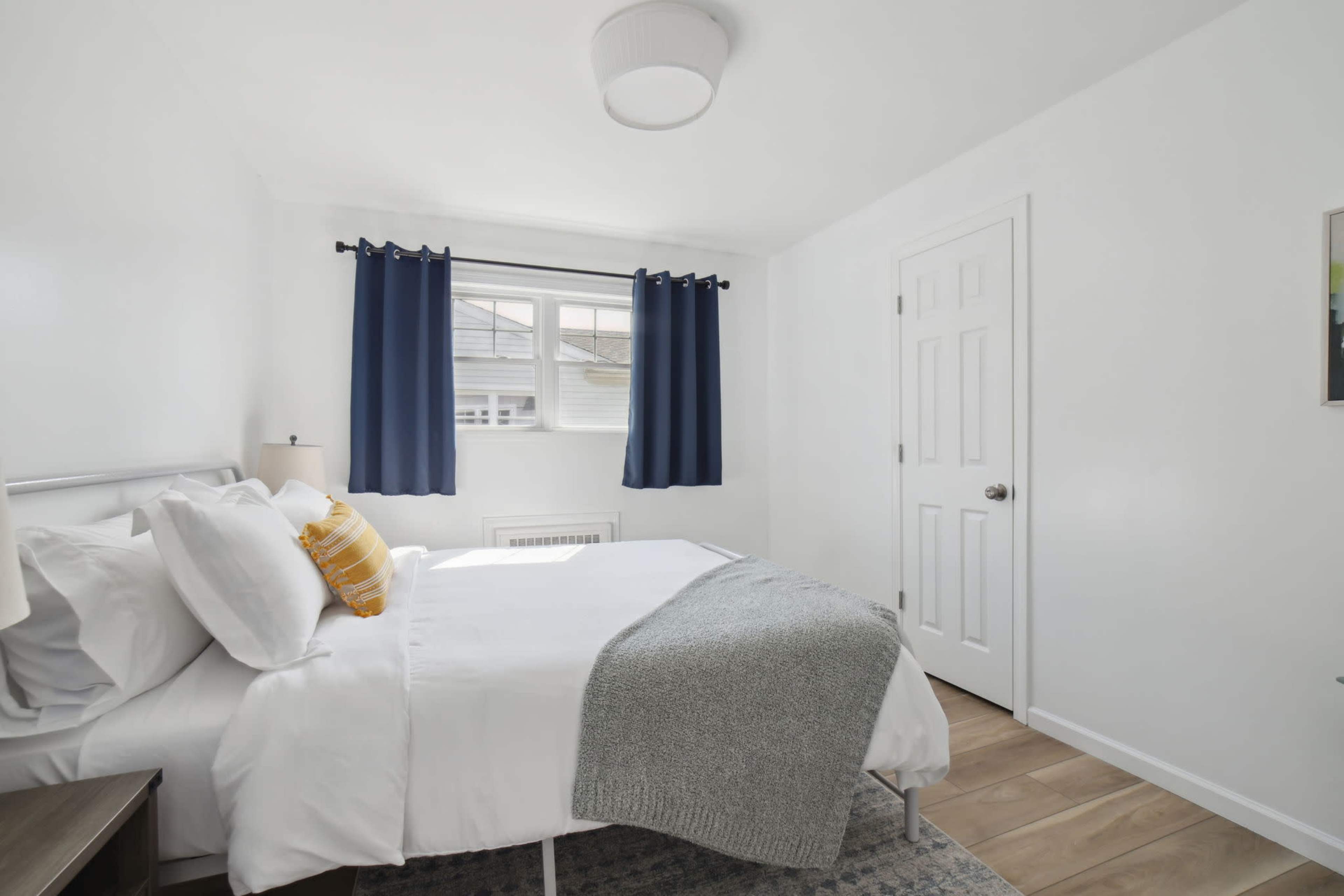 A small, neatly arranged bedroom featuring a bed with white linens, a gray throw blanket, and blue curtains over a window.