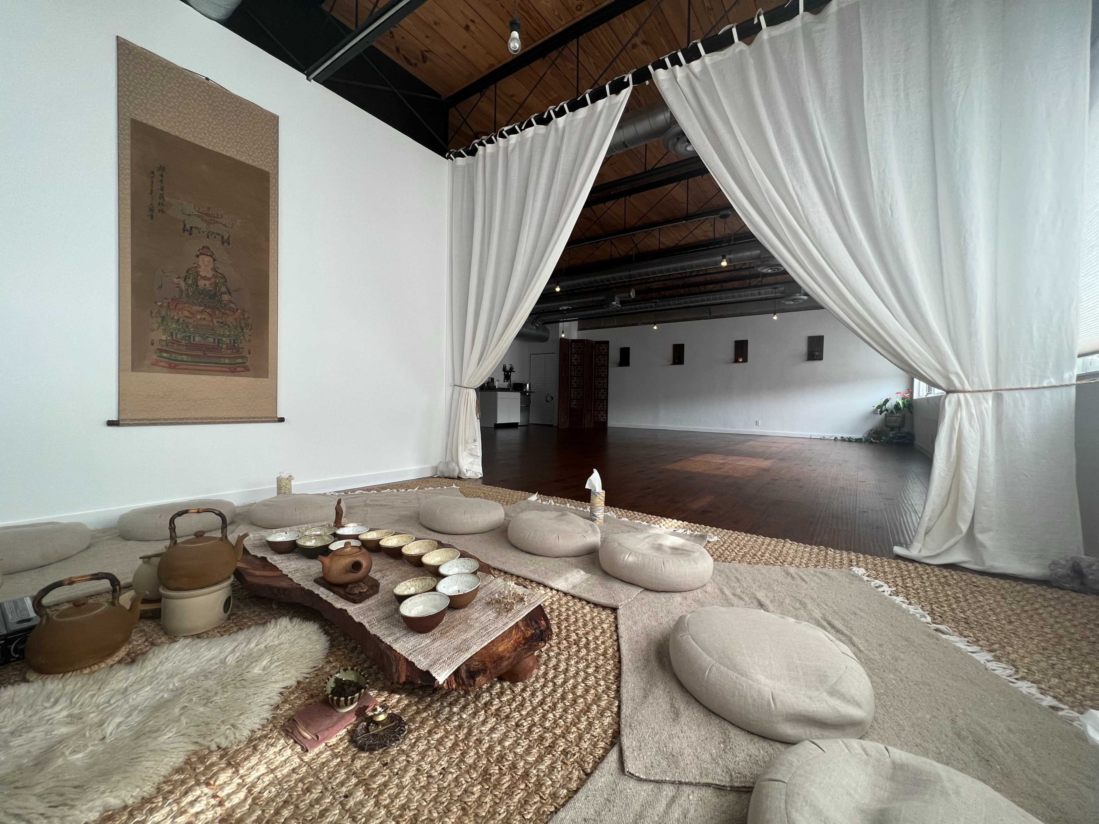 A calm interior space with floor cushions, a low wooden table set with bowls, and sheer curtains partially dividing the room.