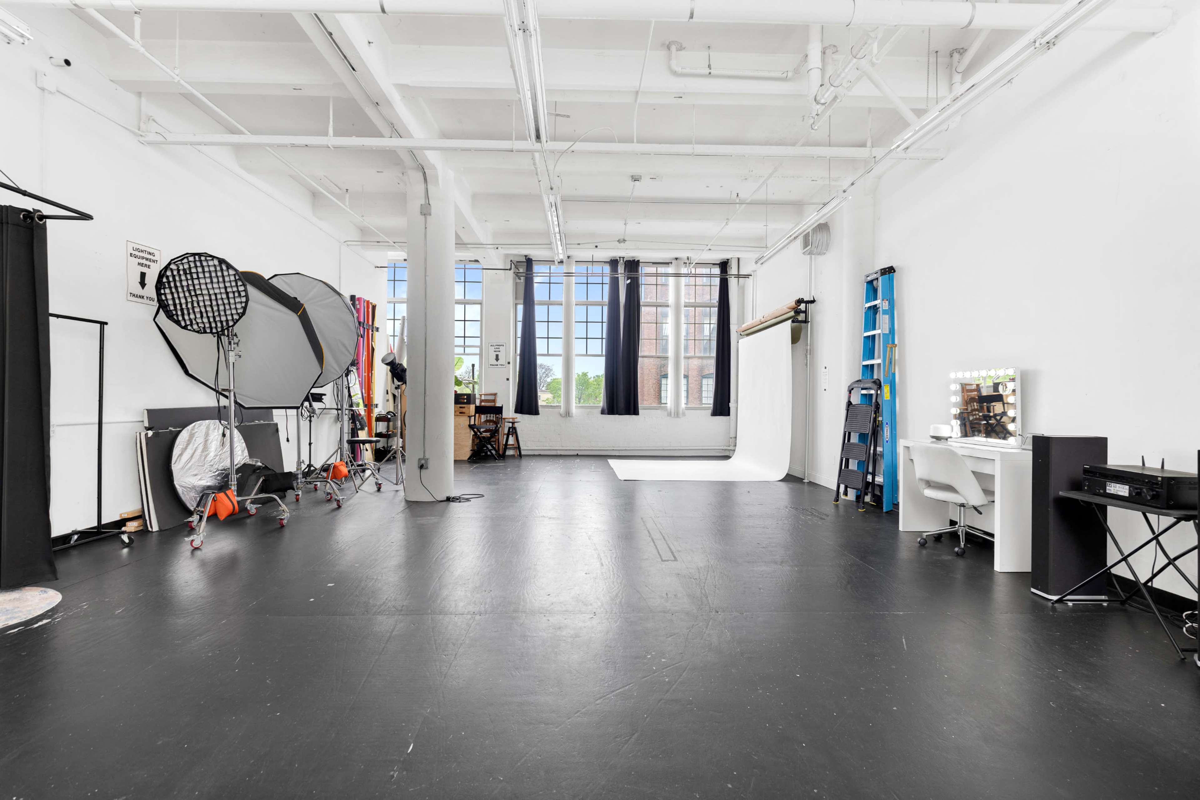 The image shows a spacious photography studio with a large open area, various equipment, and natural light streaming through large windows.