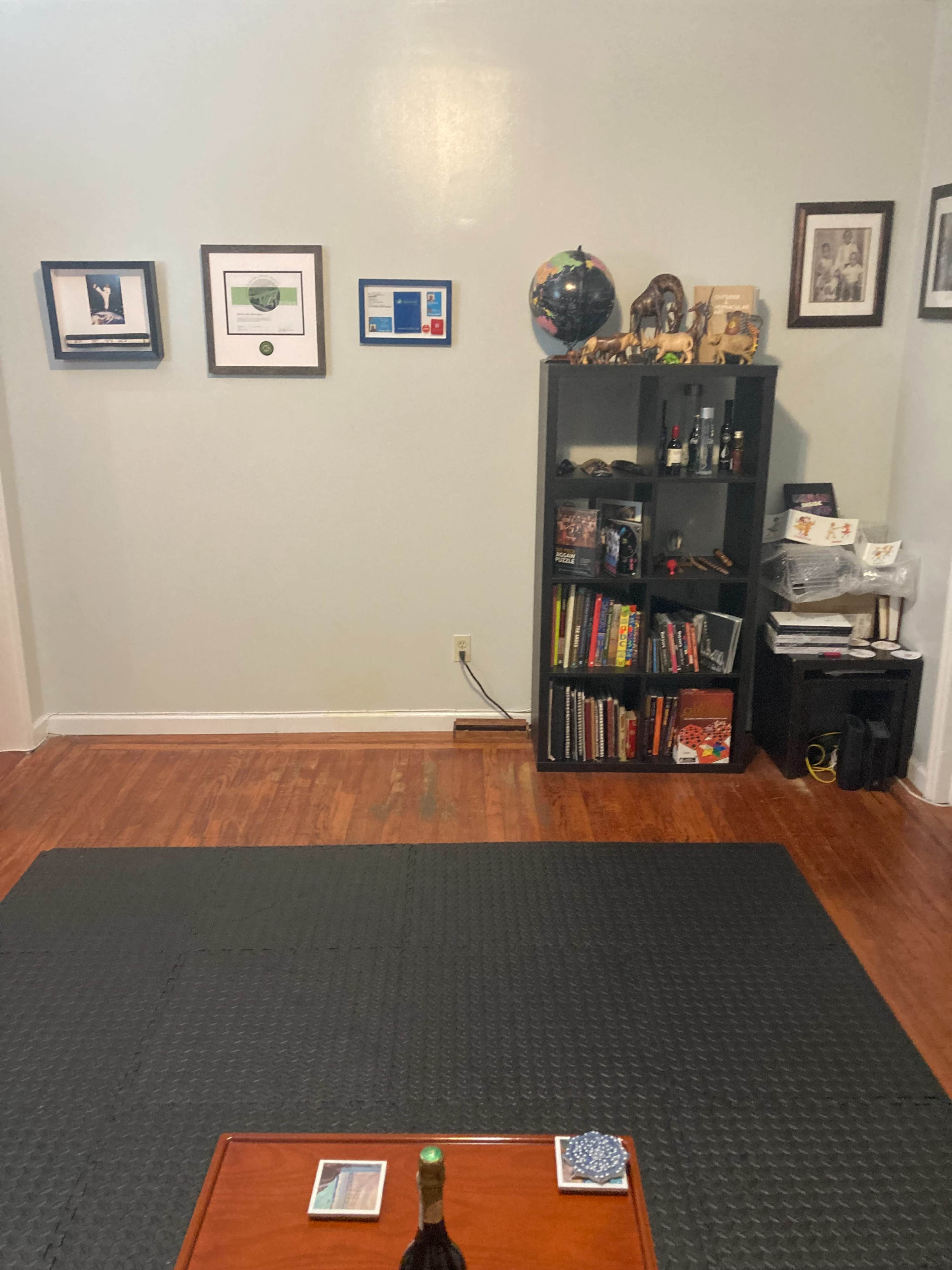 The image shows a room with a wooden floor, featuring a black rug, a low wooden table, a bookshelf filled with books and decorative items, and framed pictures on the wall.