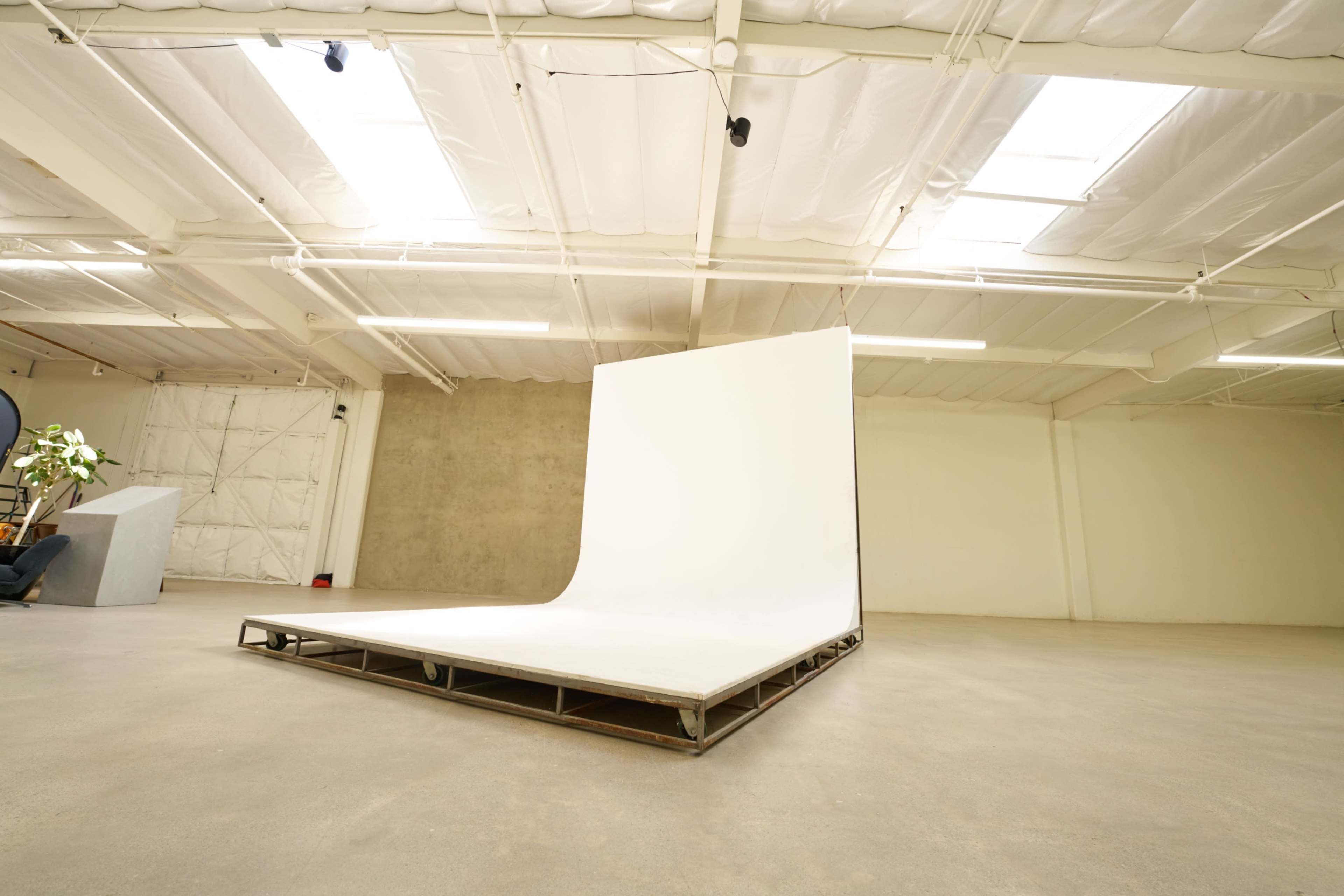 The image shows an empty photography studio with a white backdrop on a platform and large, bright windows above.