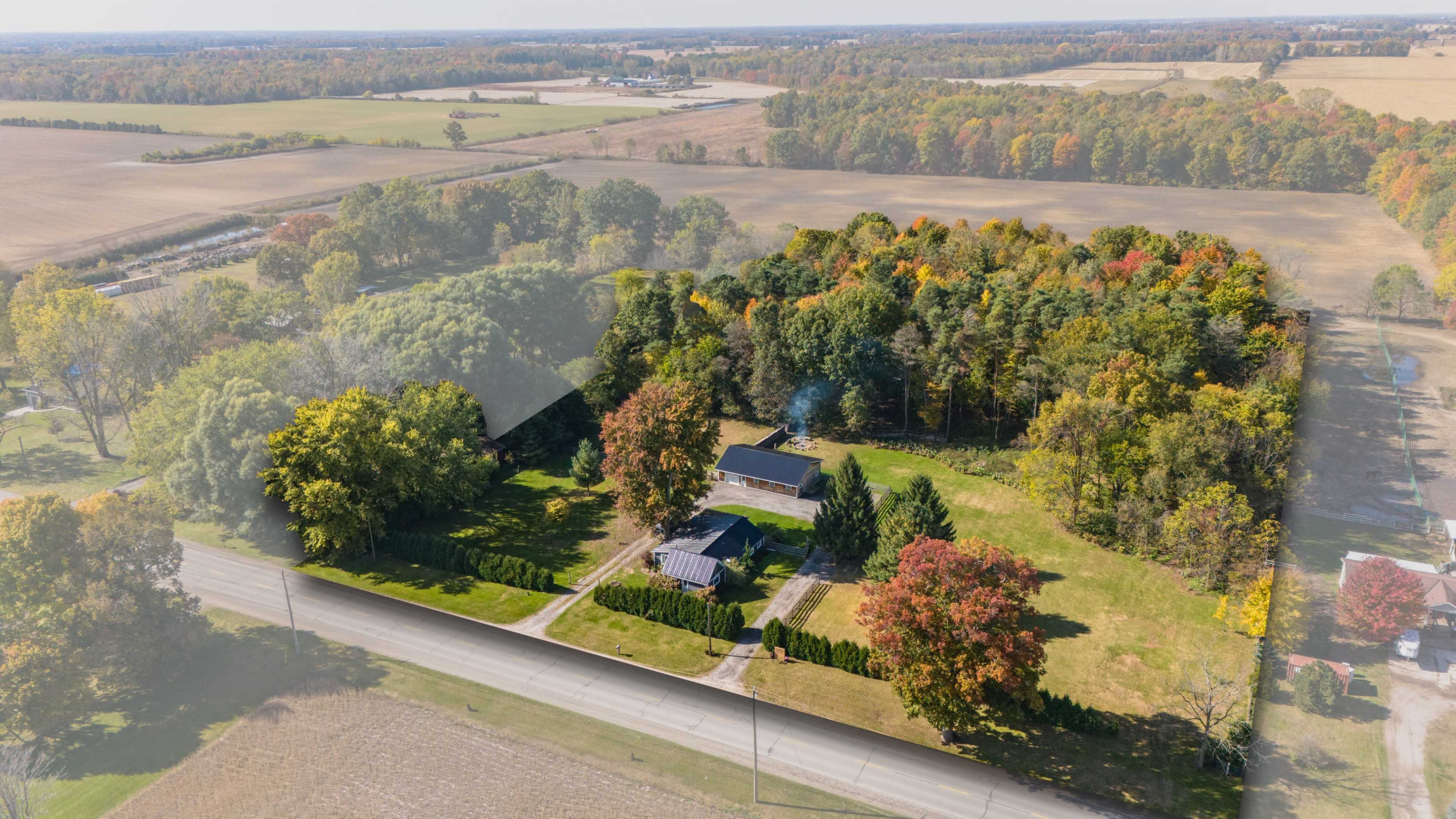 Aerial view of a house surrounded by trees displaying autumn colors, with fields extending into the distance.