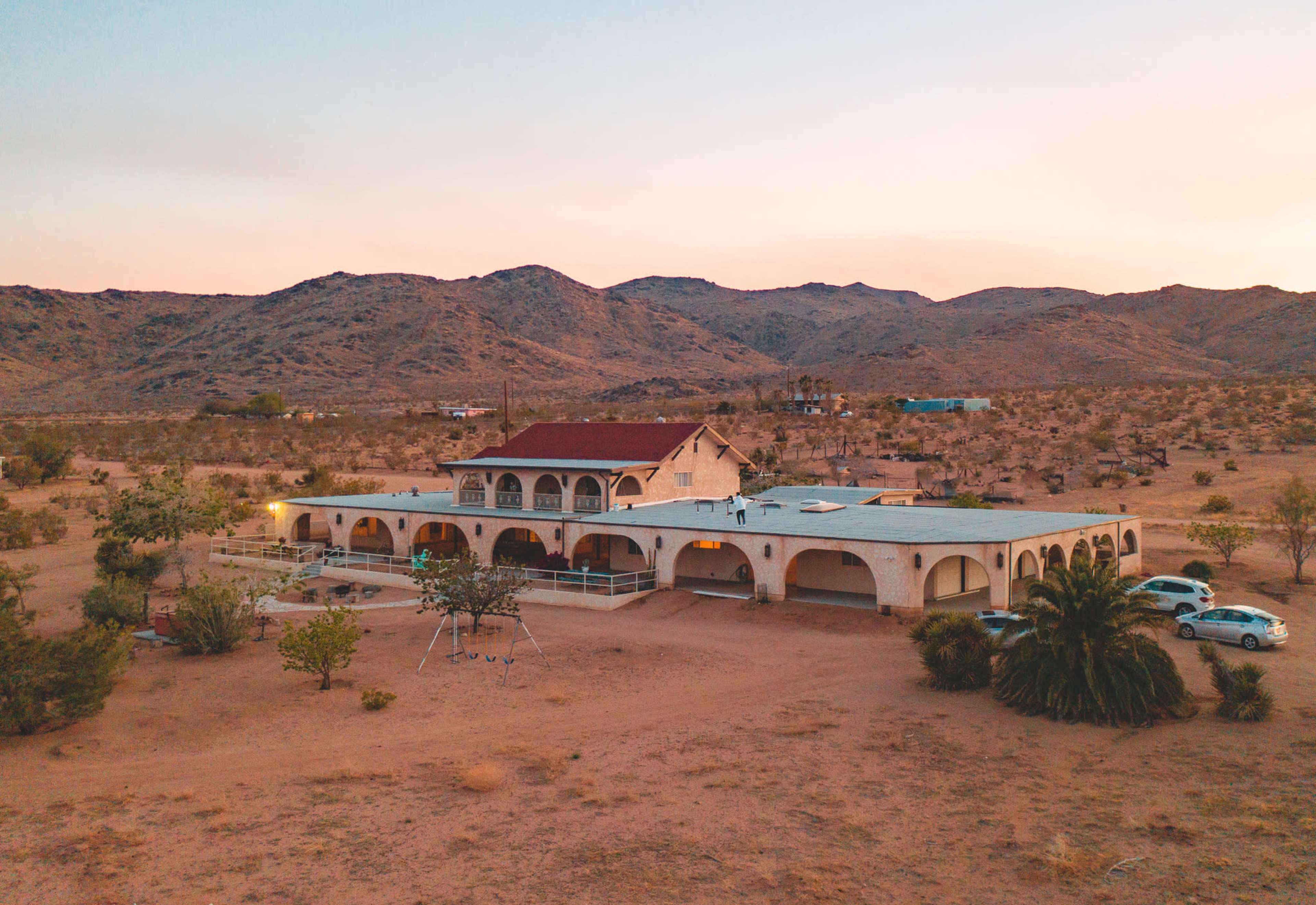 The image shows a large, multi-level house with arched features situated in a desert landscape surrounded by mountains.