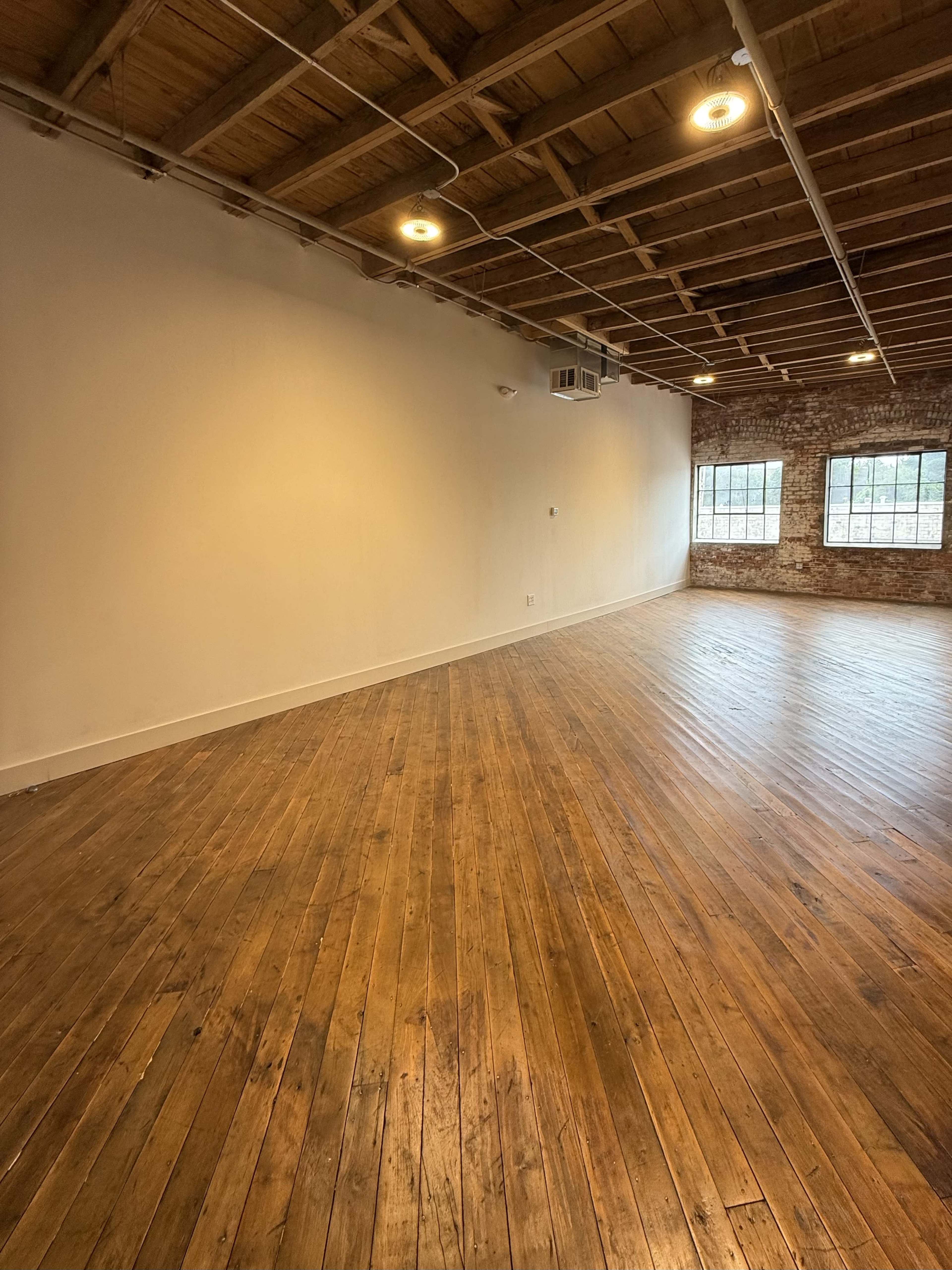 The image shows a spacious room with wooden floors, exposed brick walls, and large windows letting in natural light.