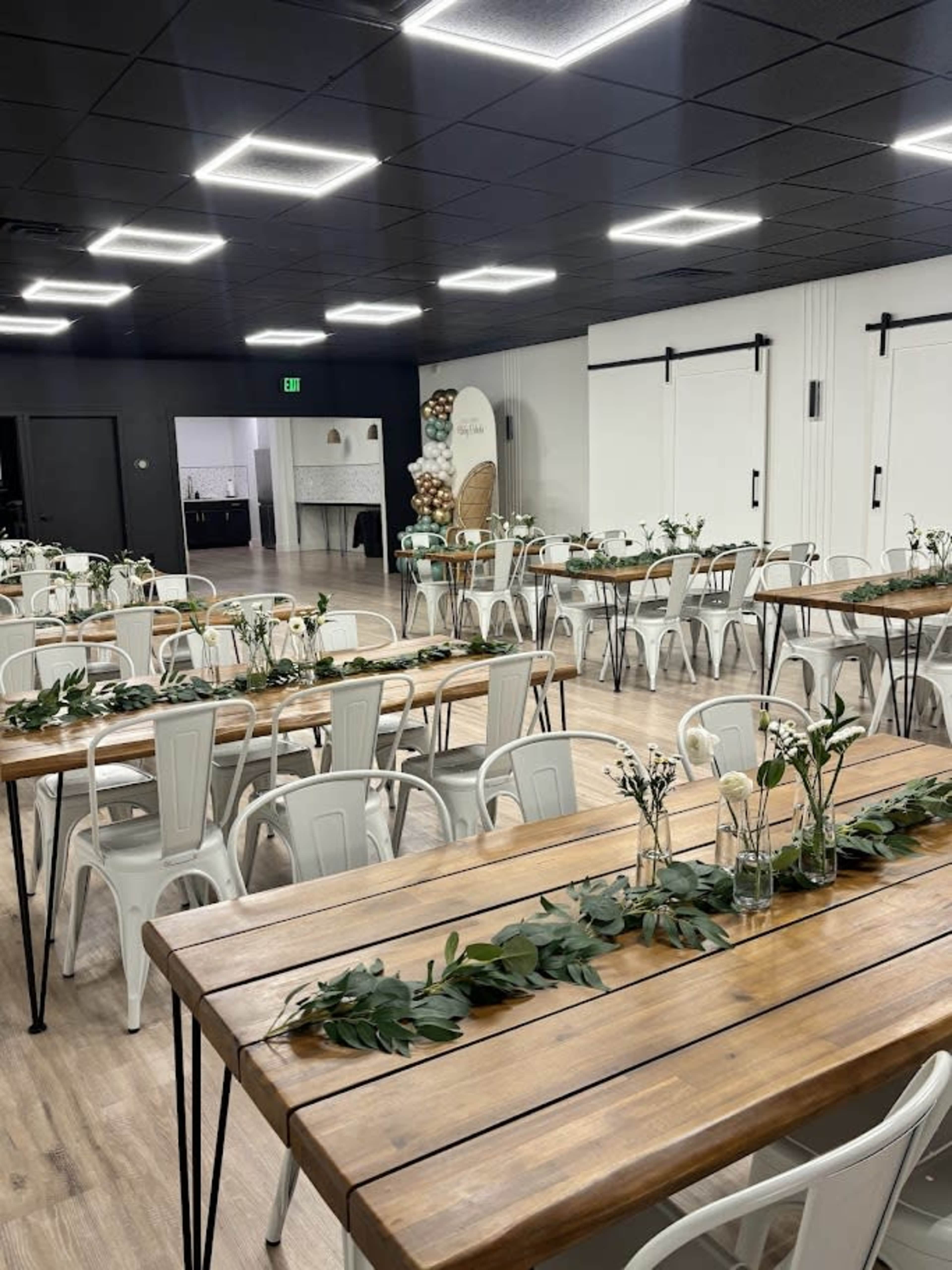 The image shows a spacious indoor venue set up with long wooden tables and white chairs, decorated with greenery and small floral arrangements.