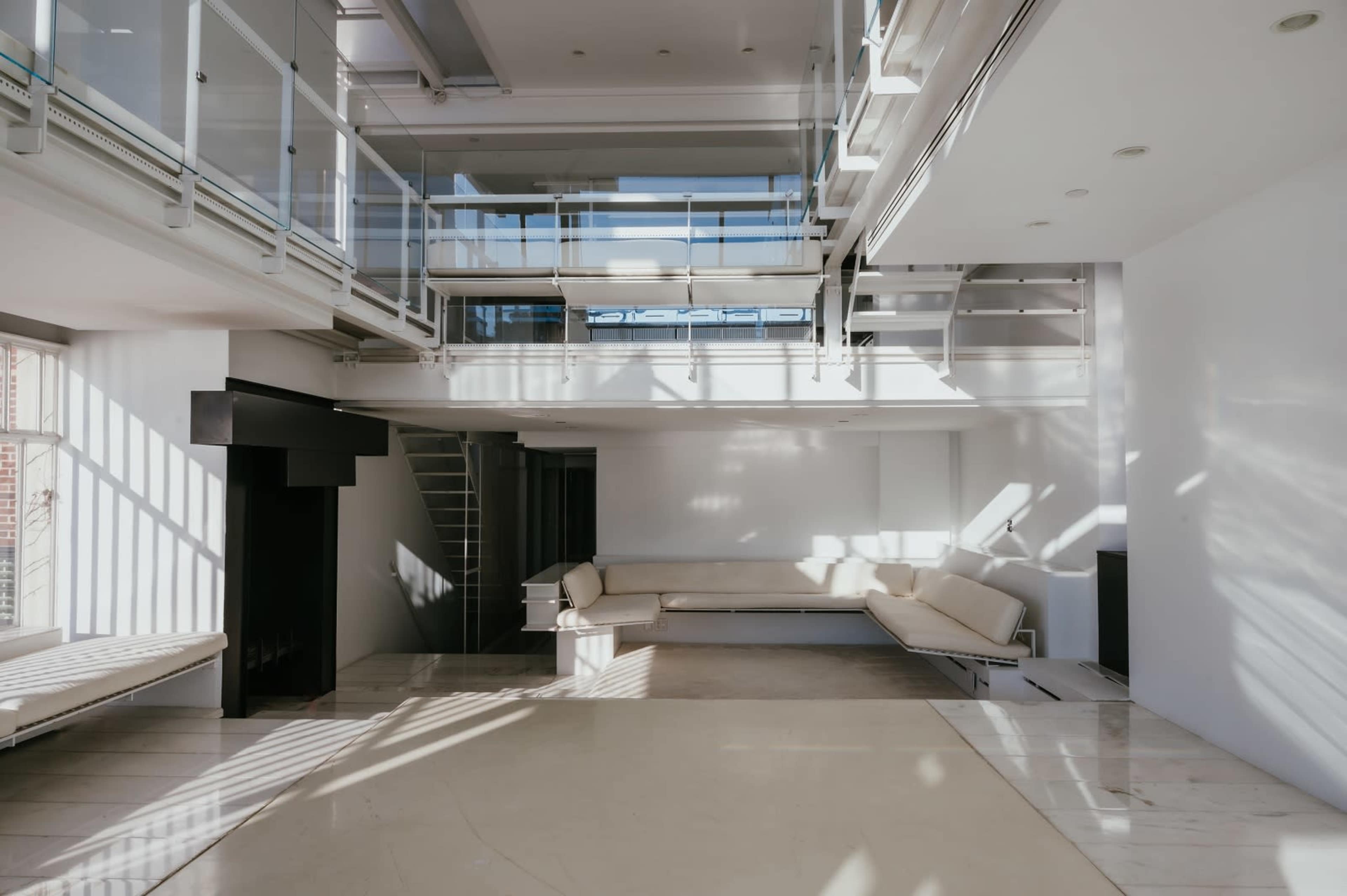 The image shows a spacious, modern interior with multiple levels, large windows allowing natural light, and minimalist furniture.