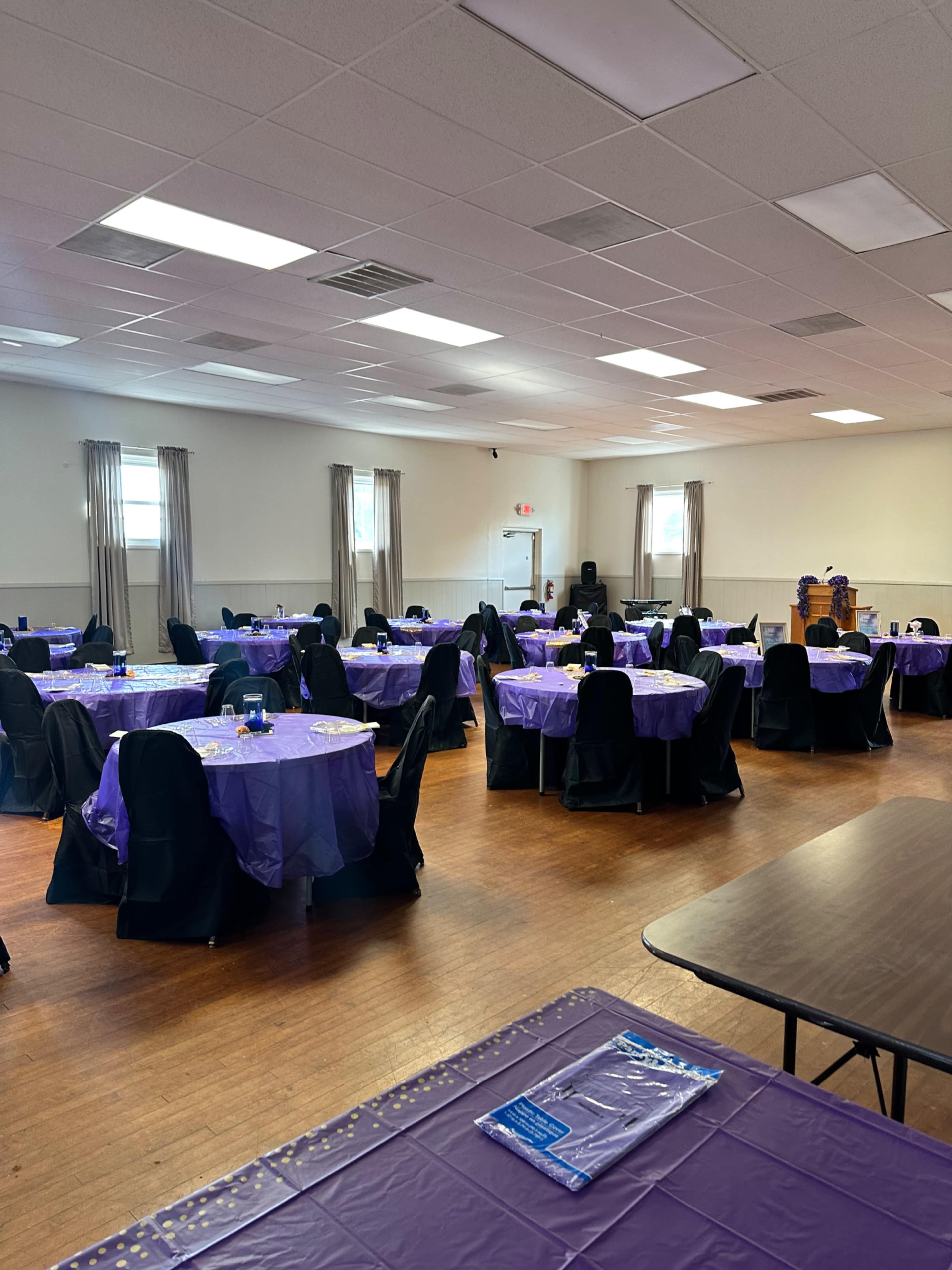 The image shows a spacious room set up for an event, with round tables covered in purple tablecloths and black chairs arranged around them.