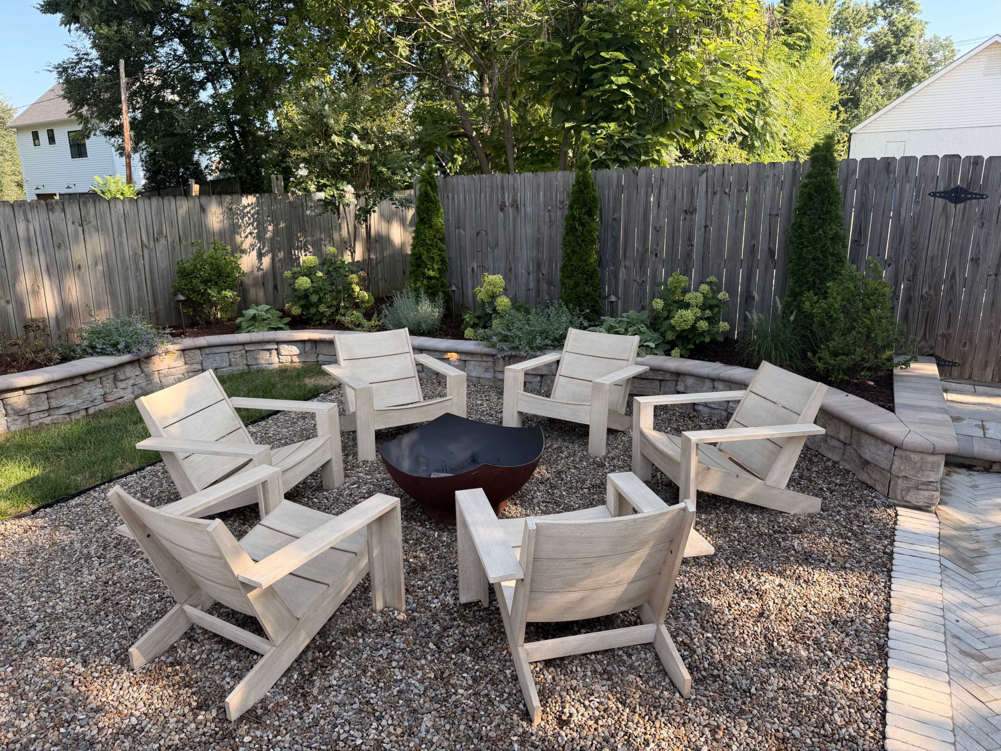 A circular arrangement of wooden chairs surrounds a fire pit in a gravel outdoor space, bordered by plants and a wooden fence.