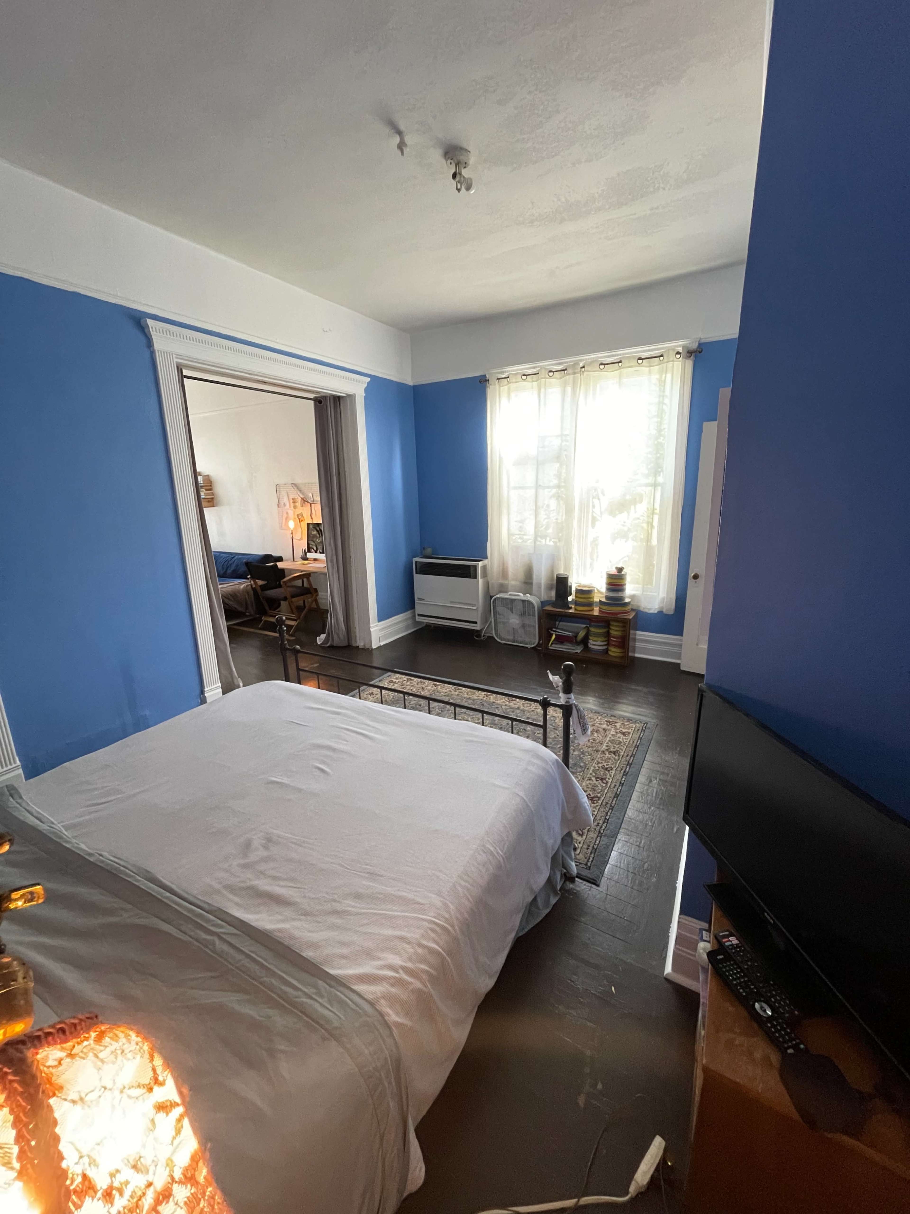 The image shows a bedroom with blue walls, a bed with a white comforter, a window with sheer curtains, and a television on a wooden cabinet.