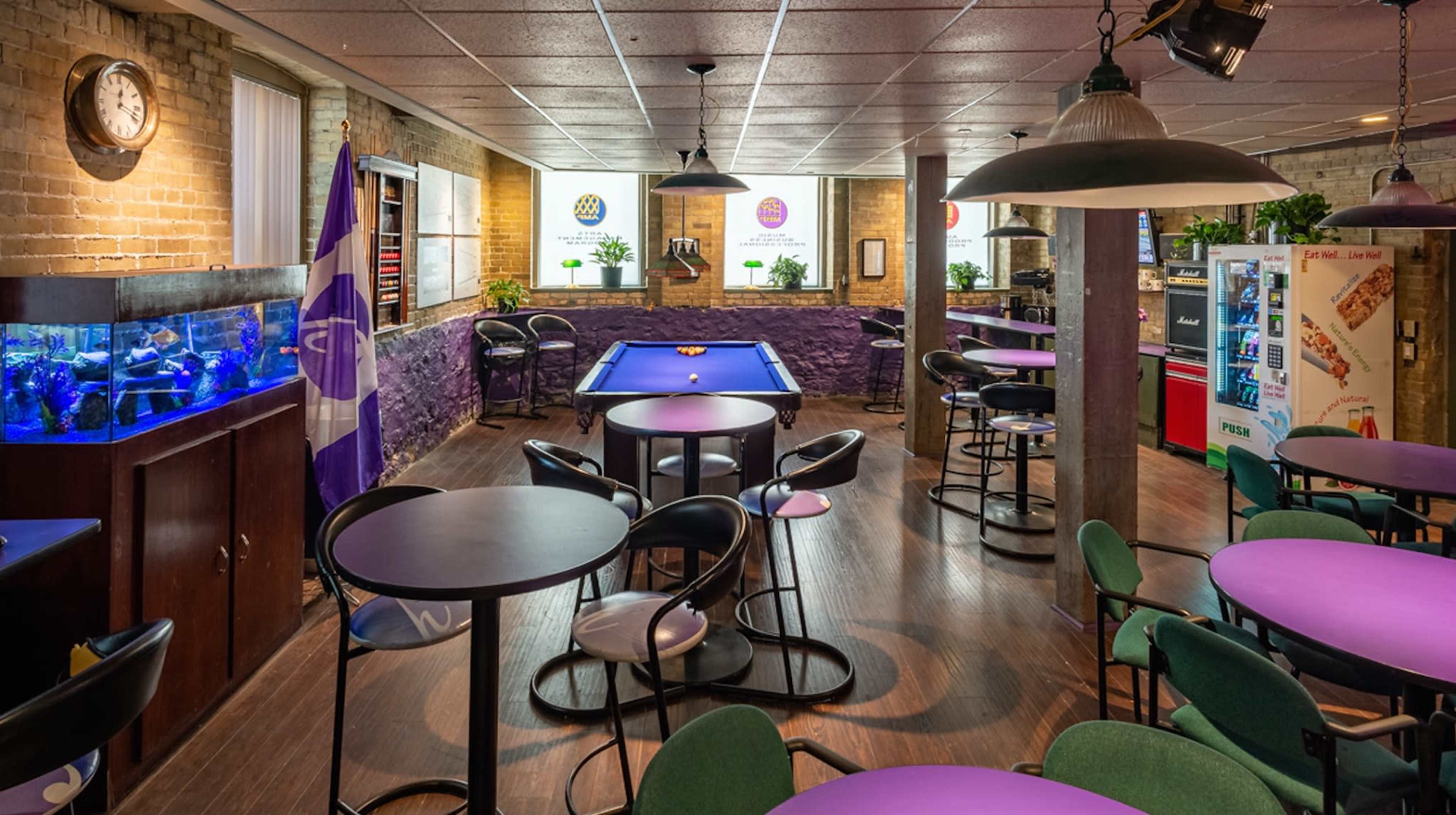 The image shows a spacious game room with a billiards table, tables and chairs, an aquarium, and vending machines against a backdrop of exposed brick walls.
