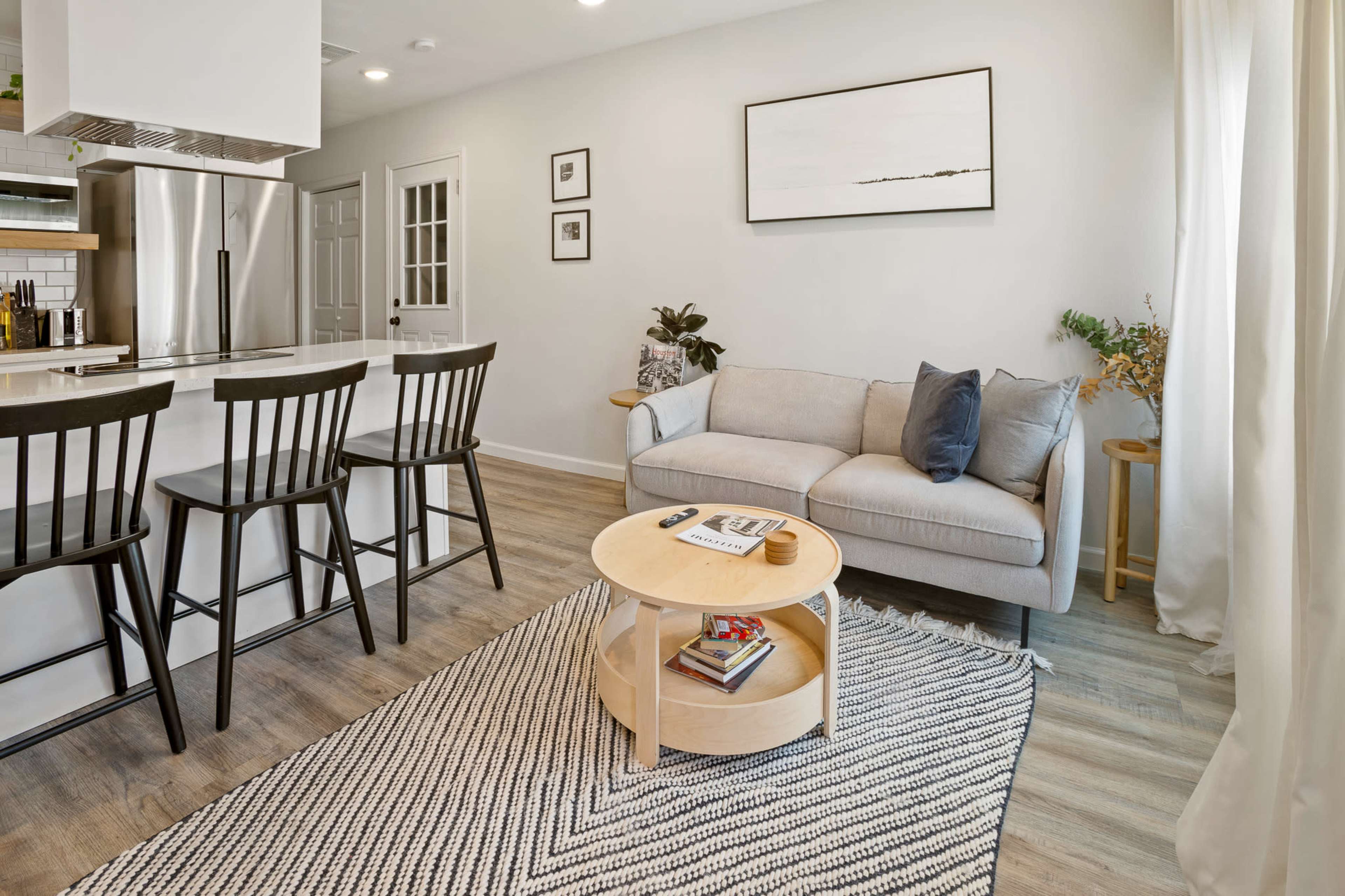 The image shows a modern living space featuring a light gray sofa, a round wooden coffee table with books, a woven rug, and a kitchen bar with three black stools.