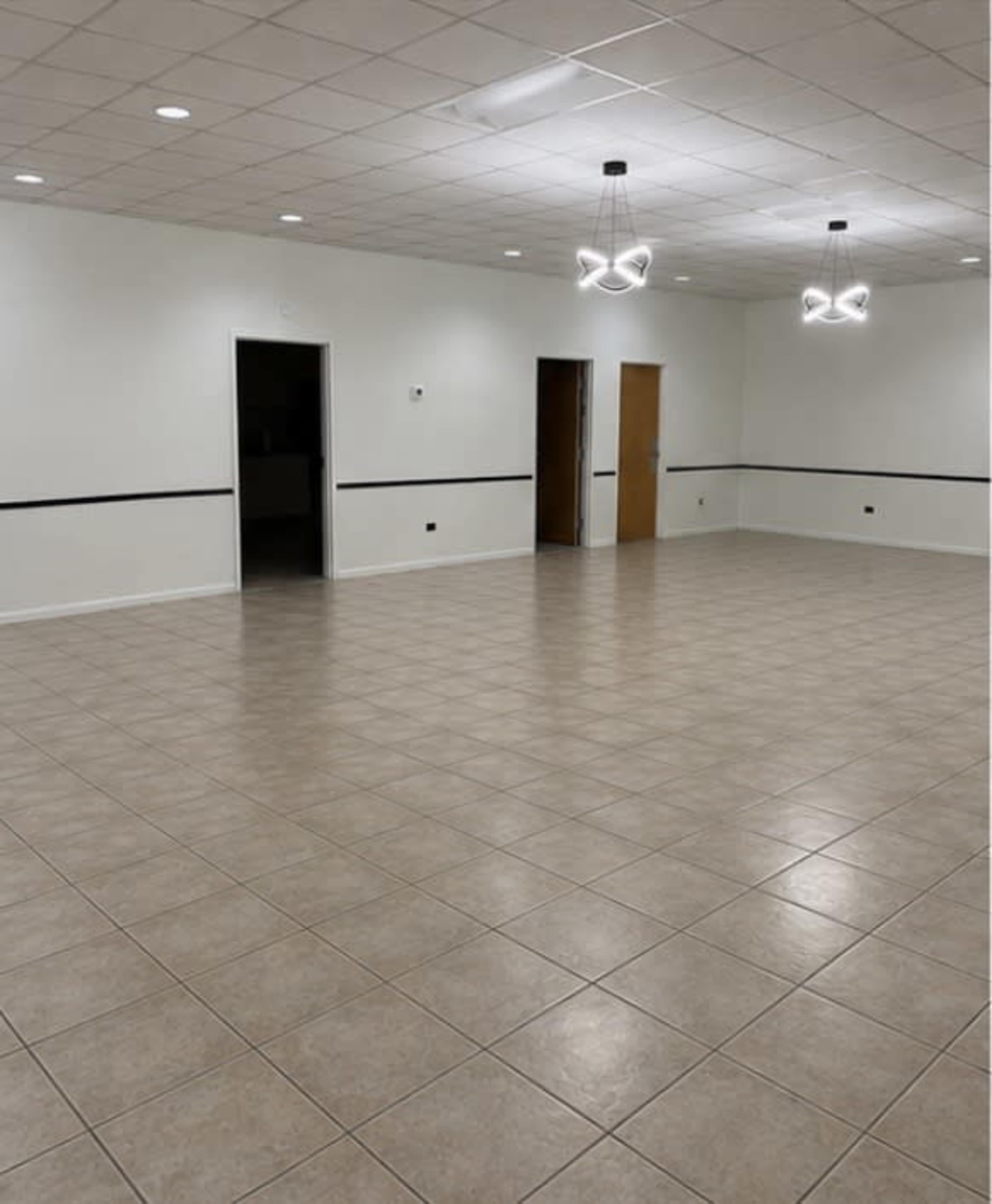 The image shows a large, empty room with beige tiled flooring, white walls, and three doors along one side, illuminated by modern light fixtures.