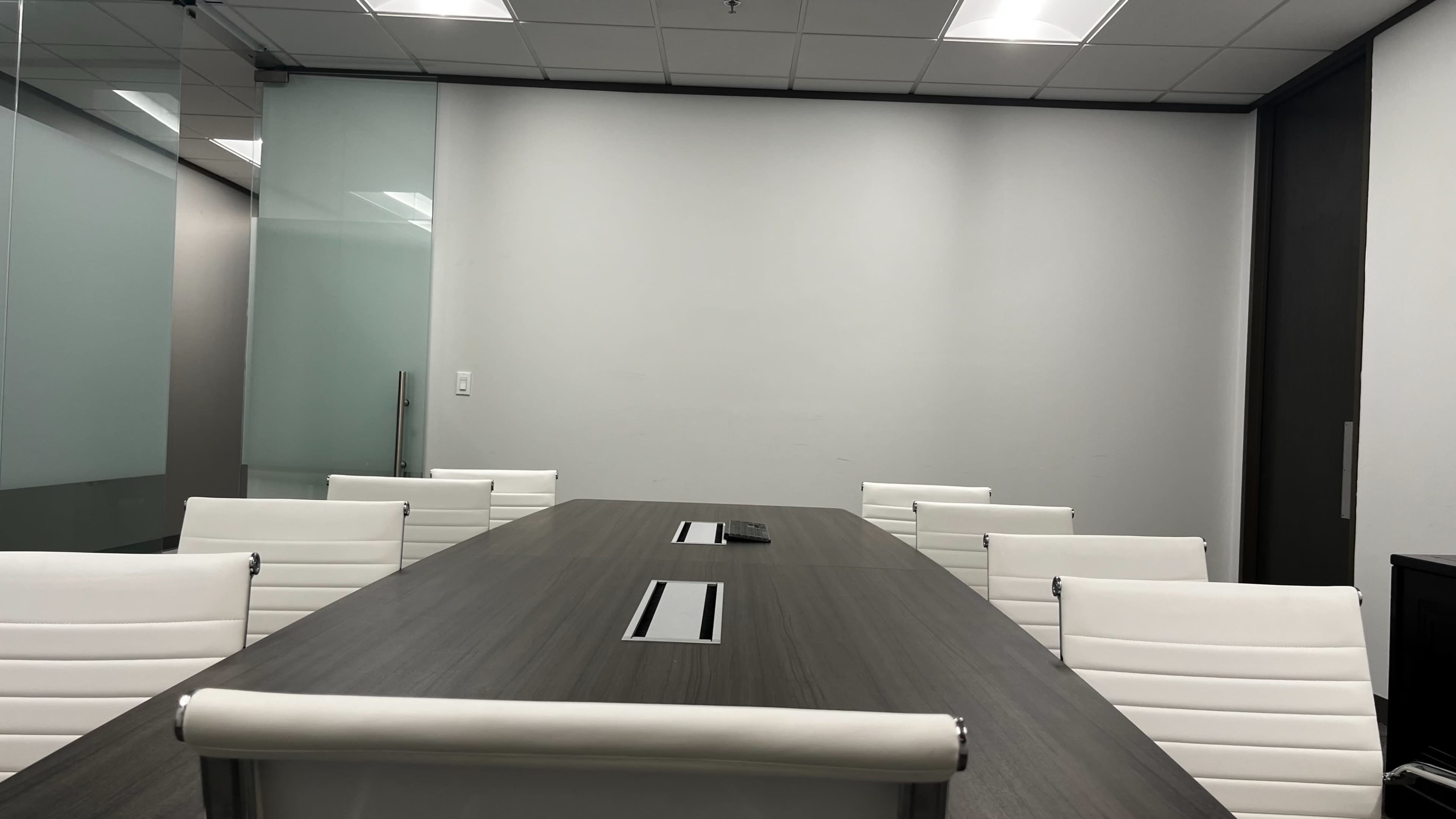 A modern conference room features a long wooden table surrounded by white chairs, with glass partitions and overhead lighting.