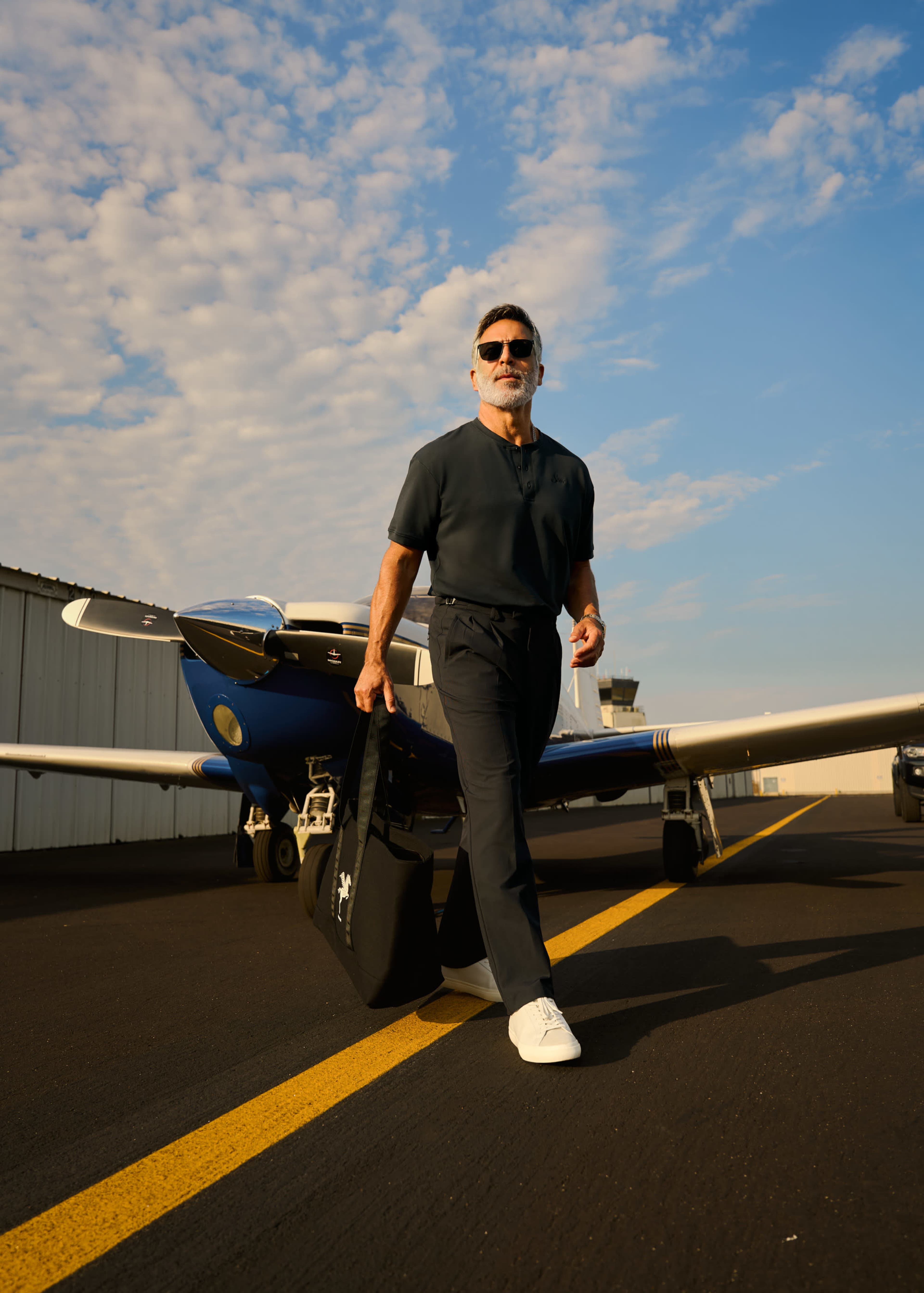 A man in a dark outfit walks towards a small airplane on an airport runway.