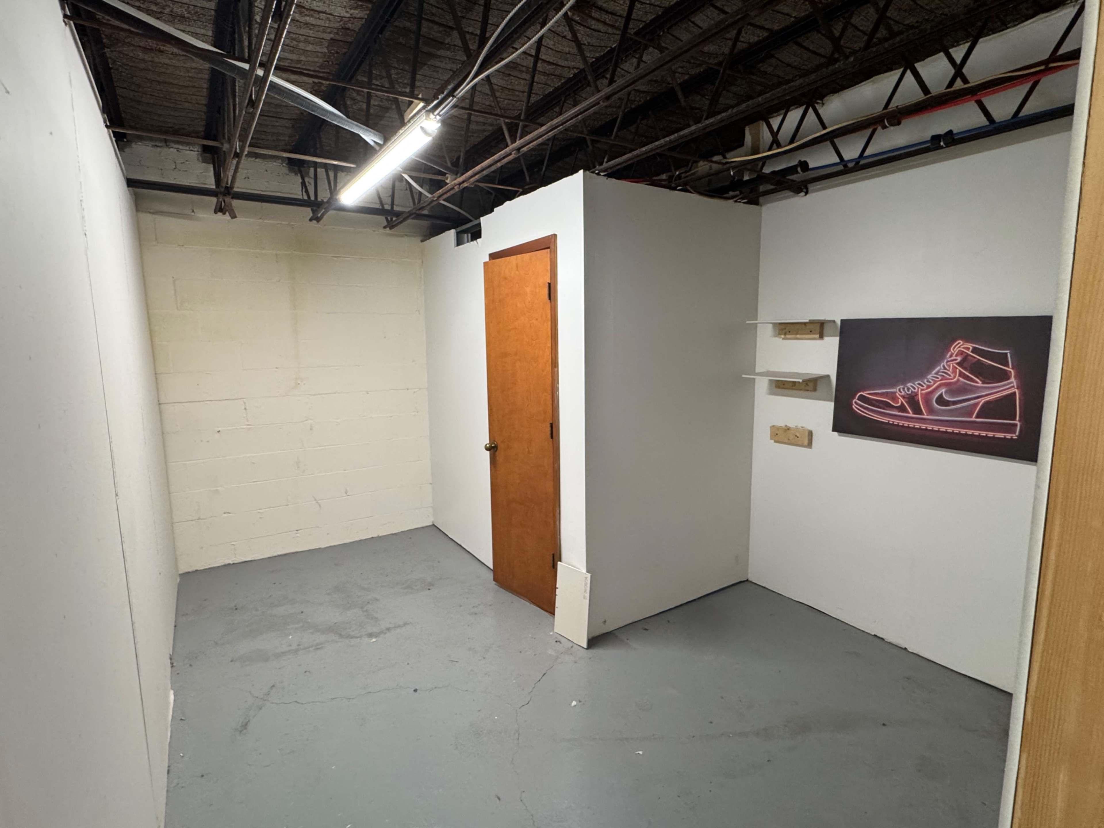 The image depicts an empty room with a concrete floor, white walls, a wooden door, and a wall-mounted shelf featuring a framed artwork of a sneaker.