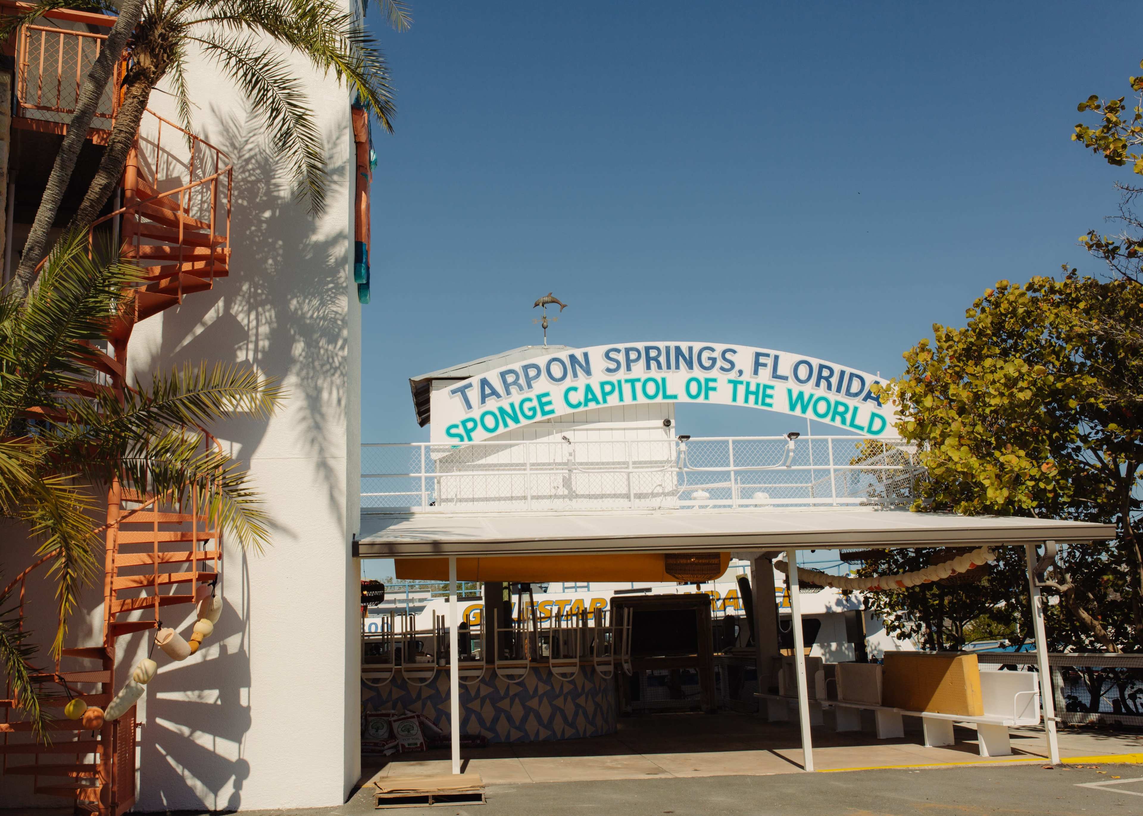 The image shows a sign reading "TARPON SPRINGS, FLORIDA SPONGE CAPITOL OF THE WORLD" above a covered area next to a white building decorated with palm trees.