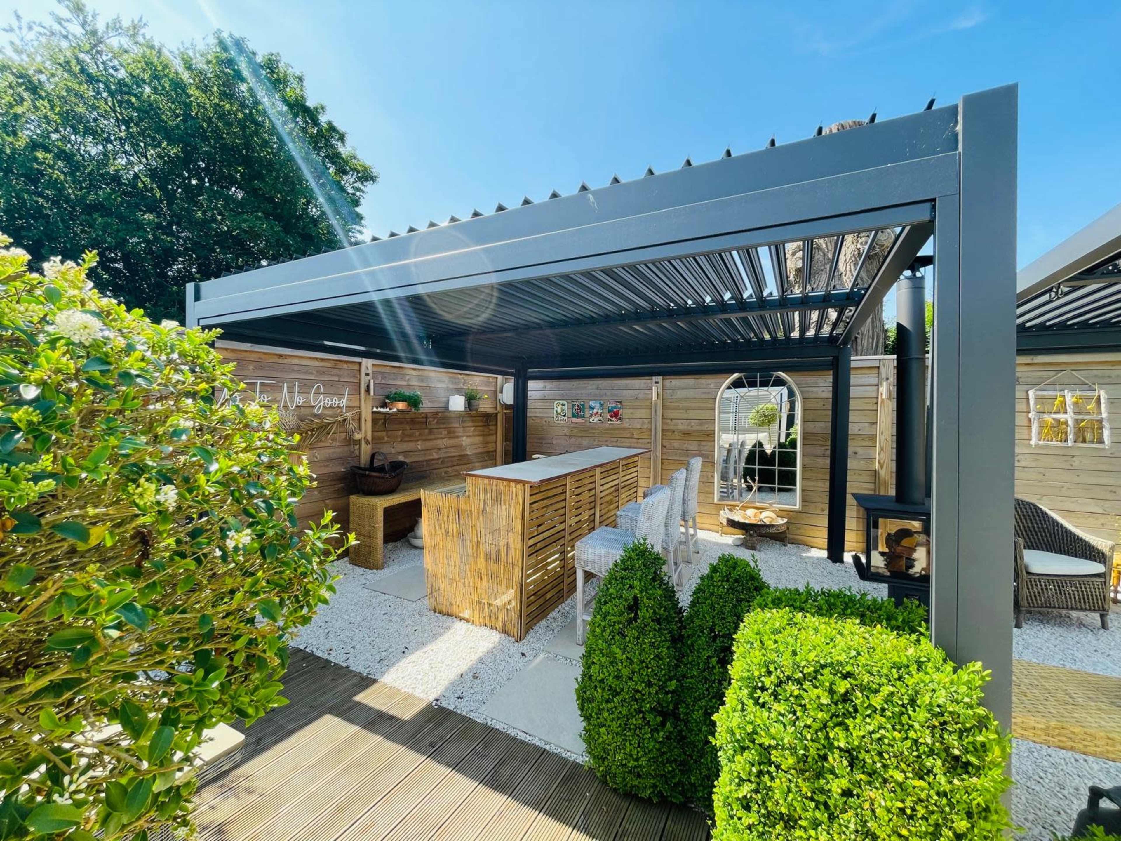 The image shows a modern outdoor dining area with a covered patio featuring a long table, chairs, and lush greenery surrounding the space.