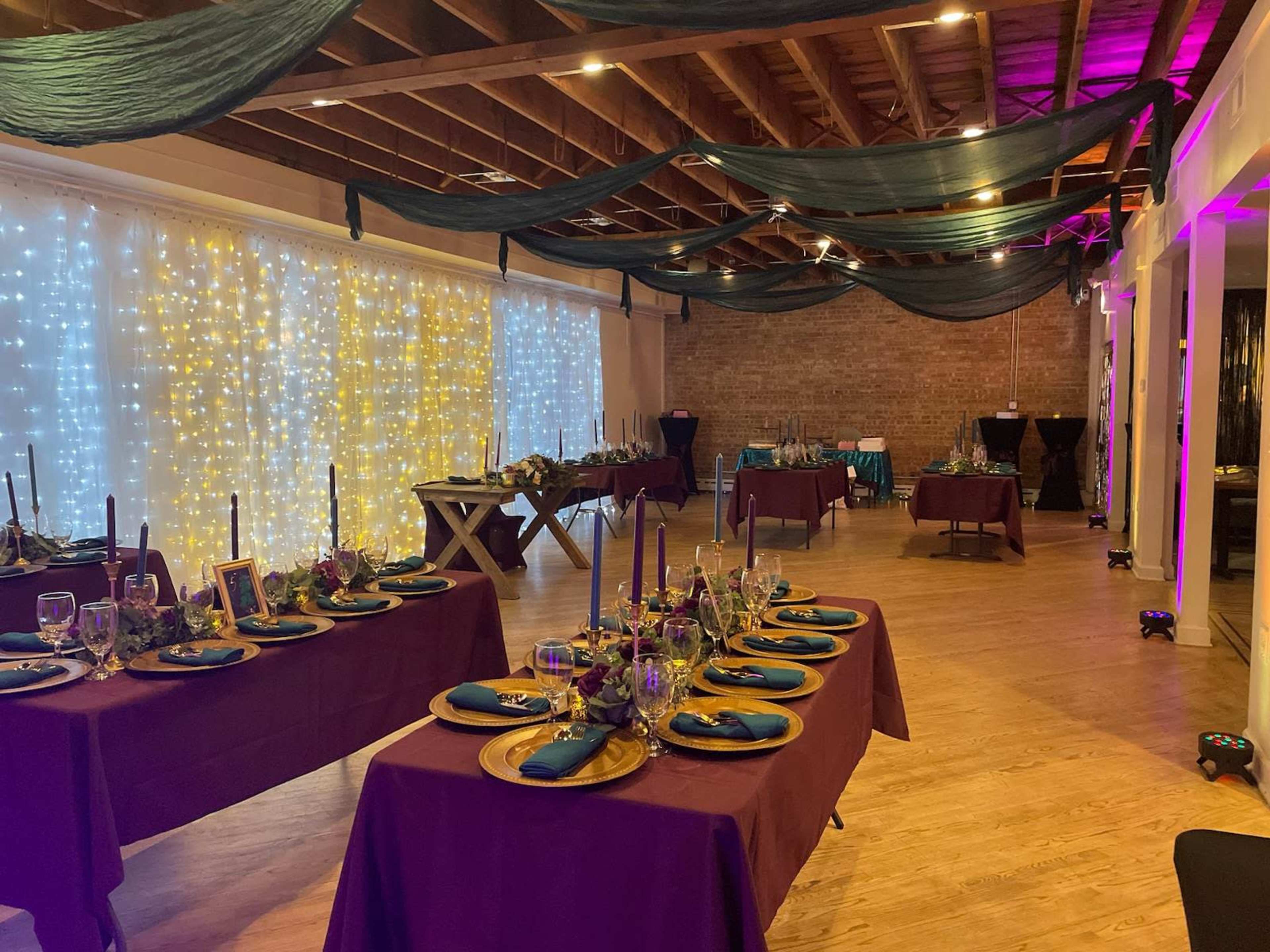 A banquet hall decorated with dim lighting, featuring tables set with blue napkins, gold plates, and floral centerpieces, against a backdrop of twinkling fairy lights.