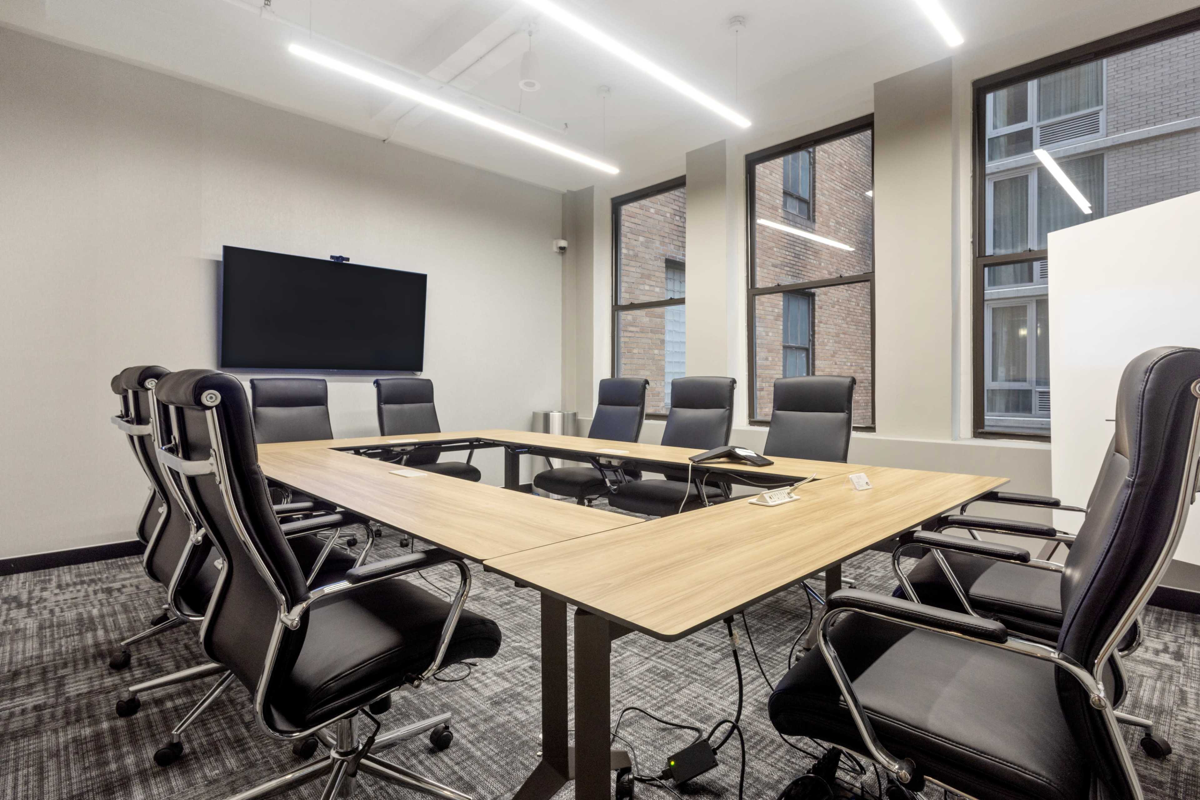 A modern conference room features a large table surrounded by ergonomic chairs, with a television screen mounted on the wall.