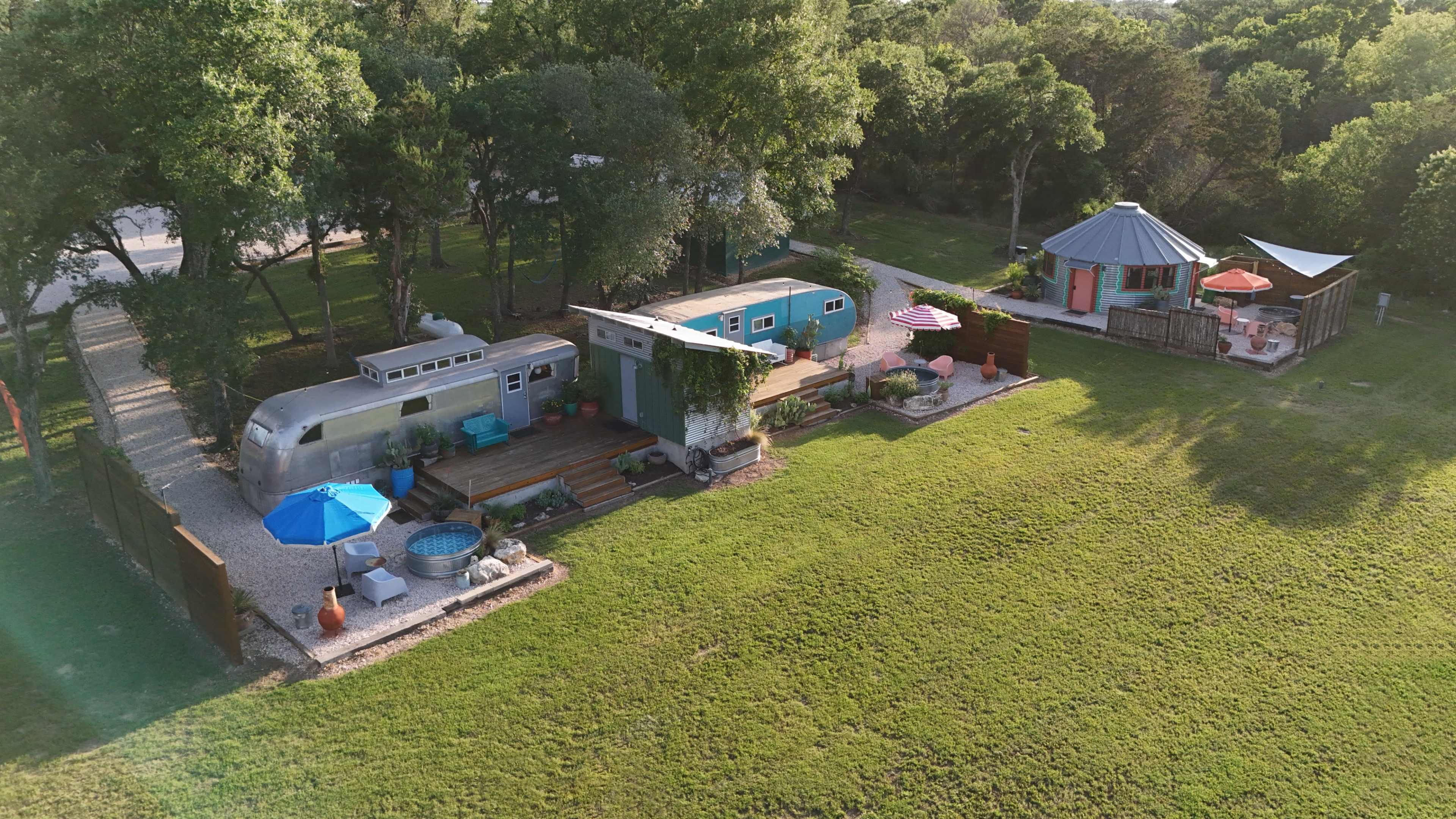 The image shows a cozy outdoor space featuring a converted Airstream trailer, a small modern building, and a circular gazebo, all set on a grassy area surrounded by trees.
