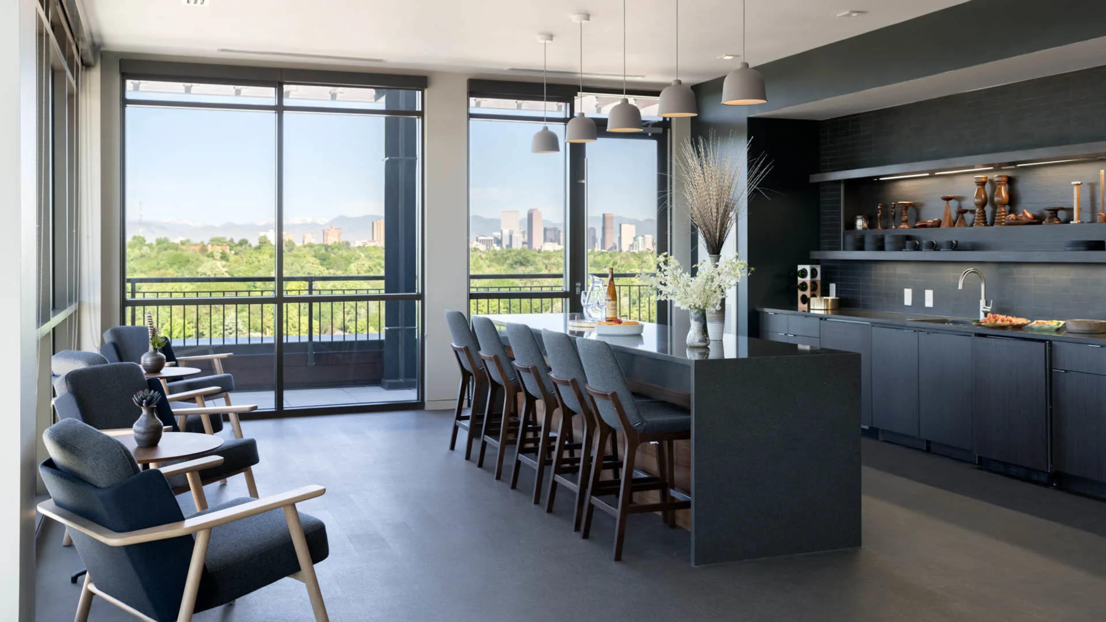 A modern kitchen and dining area with a long table and bar stools, large windows overlooking a green landscape and city skyline.