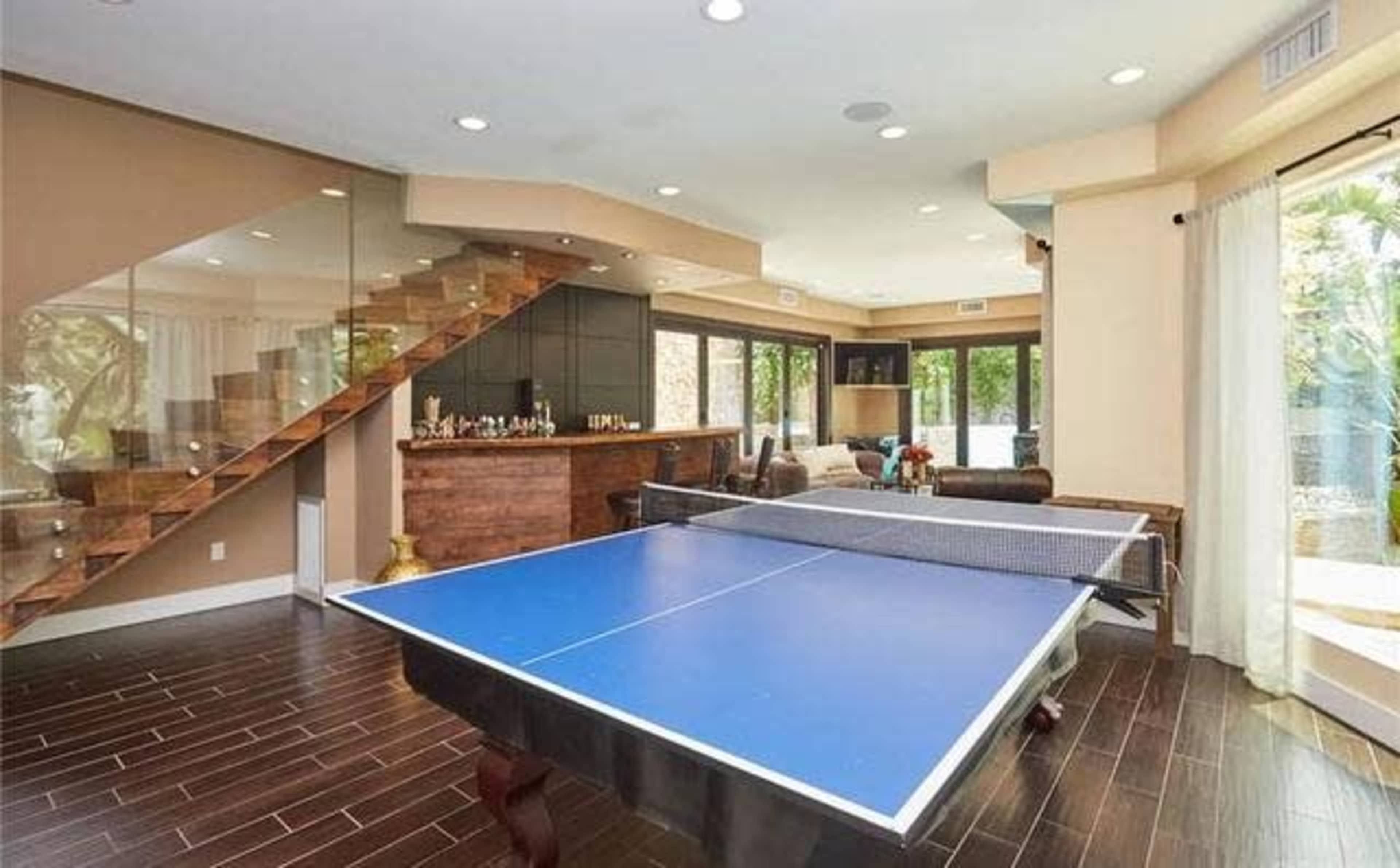The image shows a modern interior space featuring a blue ping pong table, a wooden bar area, and large windows allowing natural light.