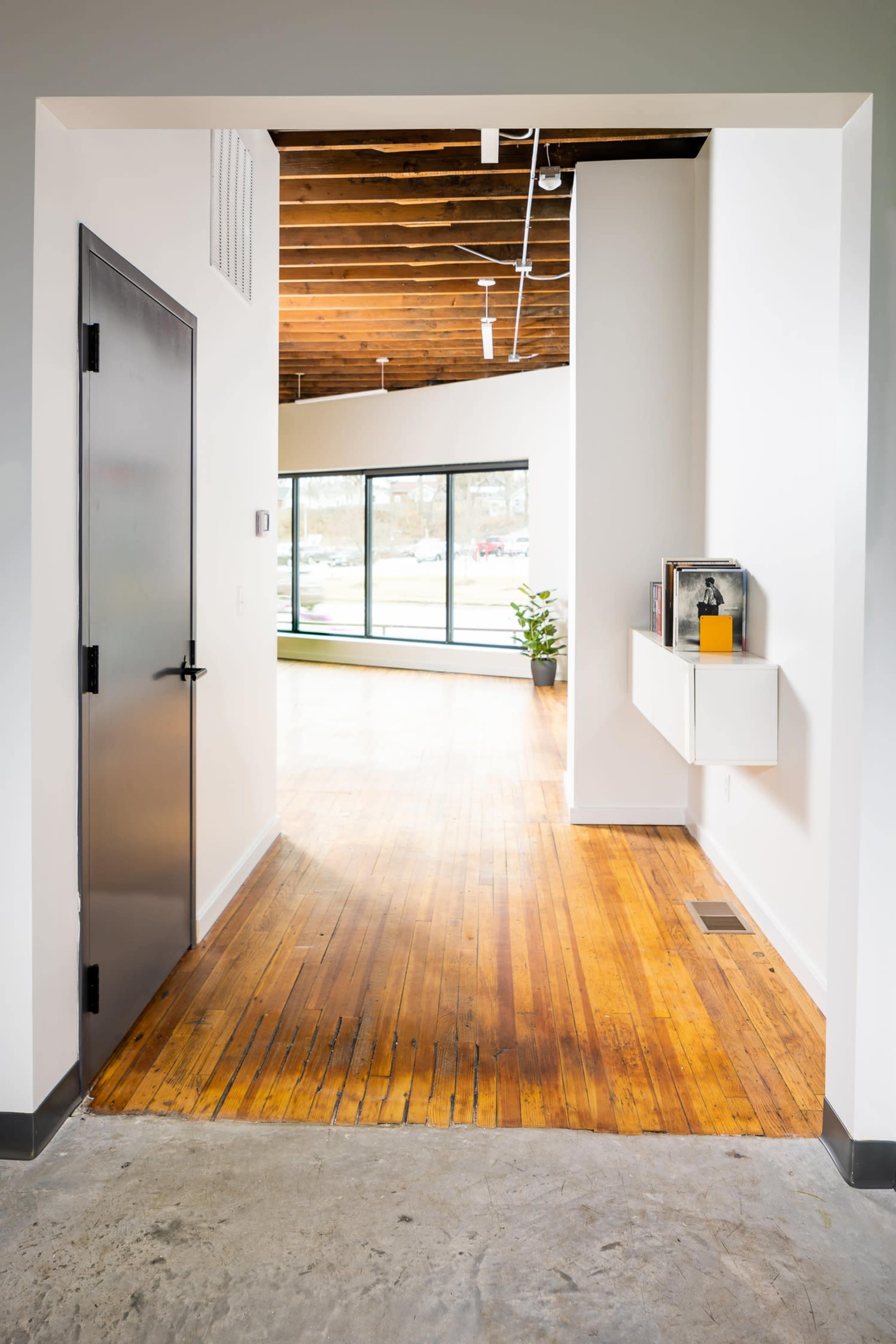 The image shows a modern interior hallway with a wooden floor leading into a well-lit room featuring large windows and minimal decor.
