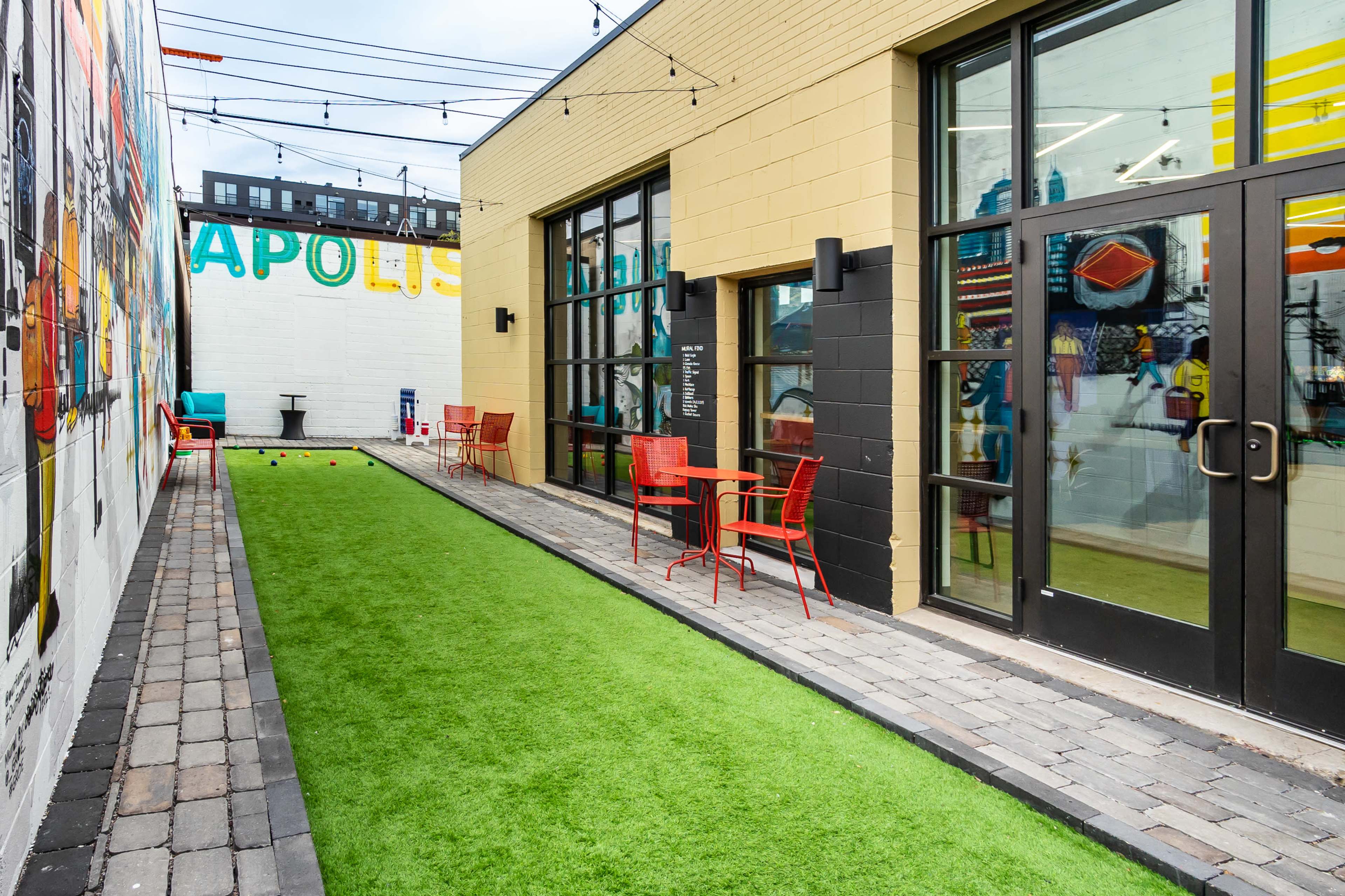 The image shows a narrow outdoor space lined with artificial grass and paved pathways, featuring a mural on one wall and red chairs and tables arranged in front of large glass doors.