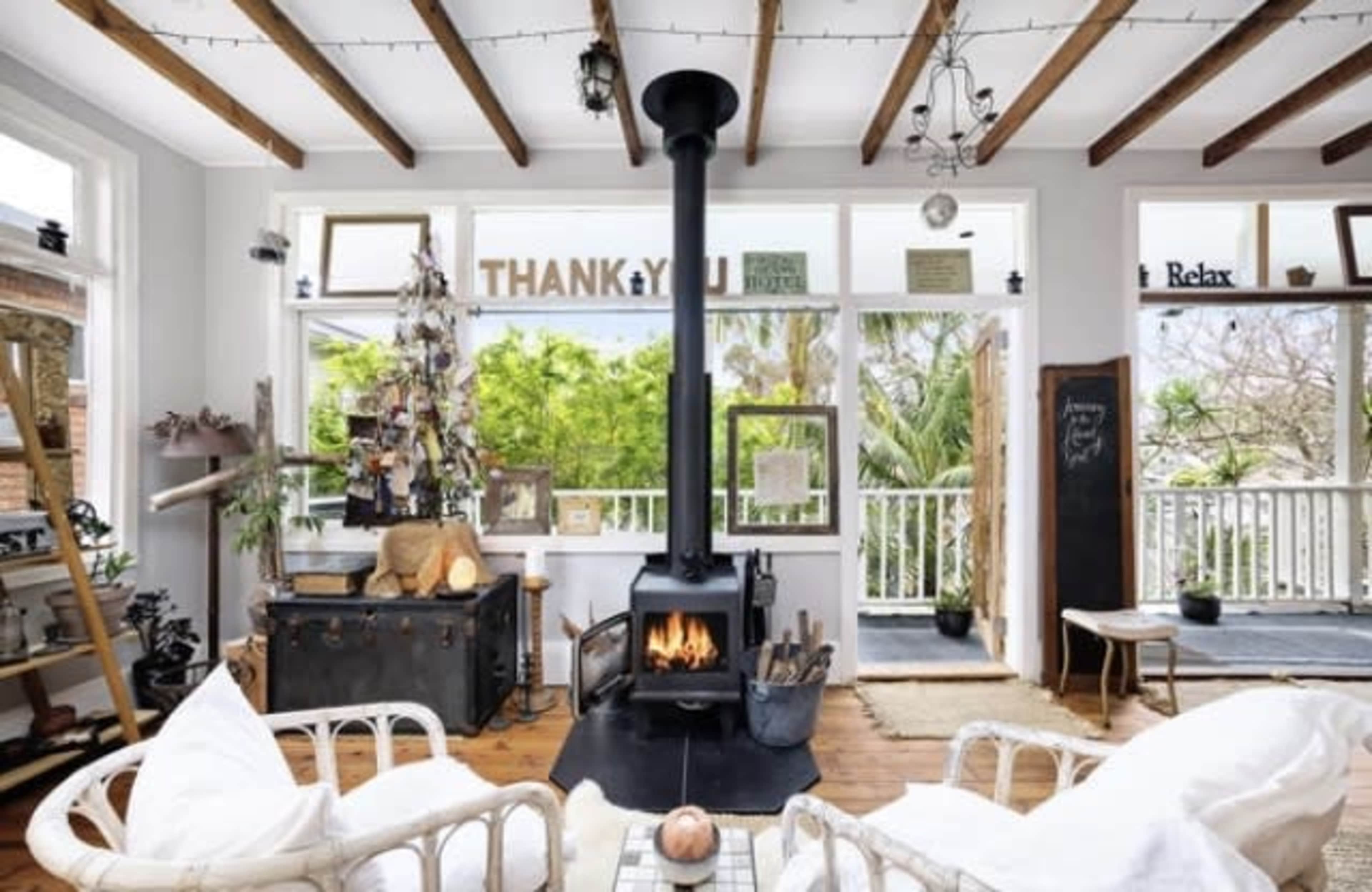 A cozy living room features a central black wood stove surrounded by light-colored furniture, large windows showcasing greenery, and wooden beams on the ceiling.