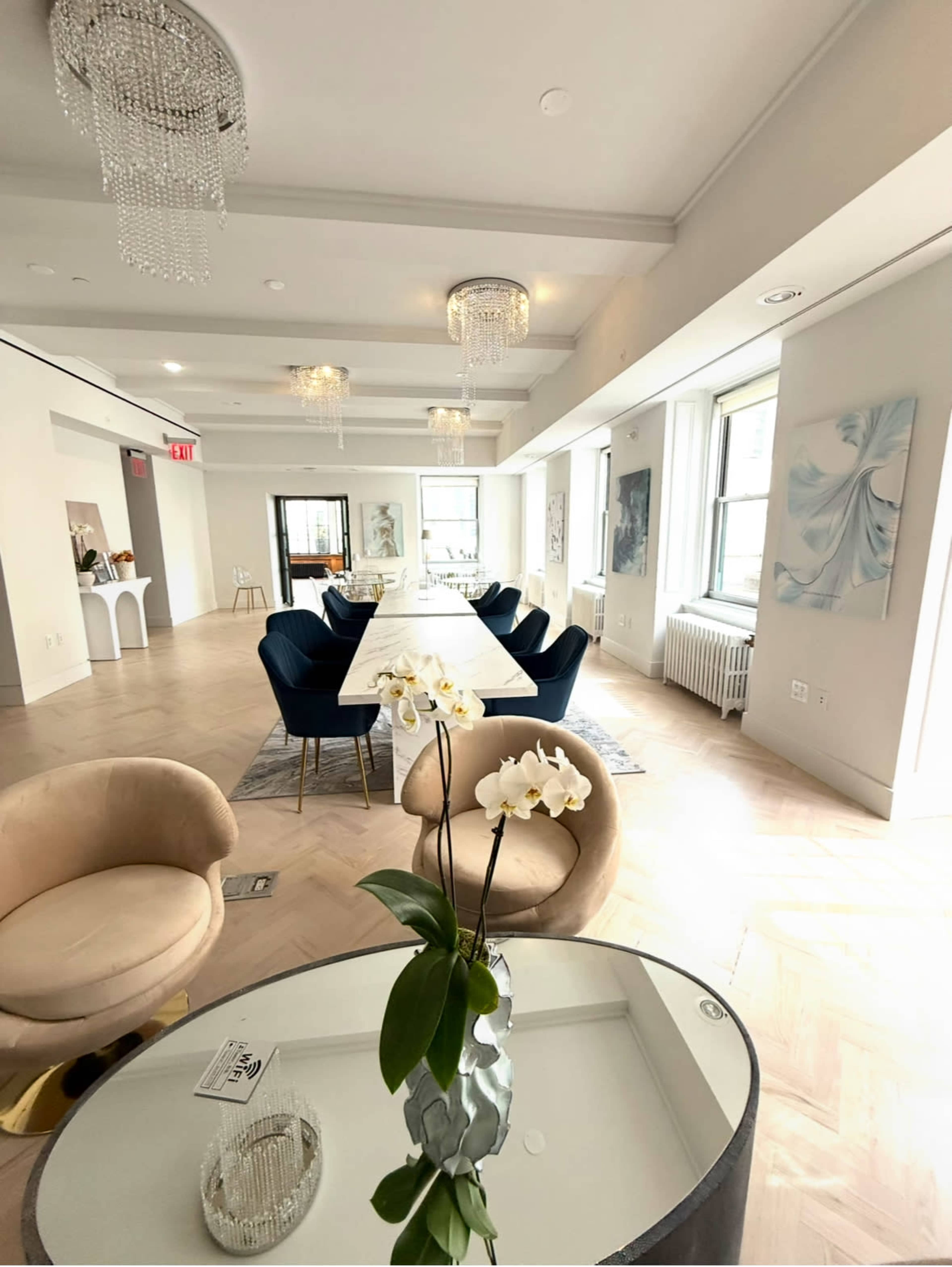 The image displays a modern dining area with a long white table surrounded by dark blue chairs, illuminated by crystal chandeliers.