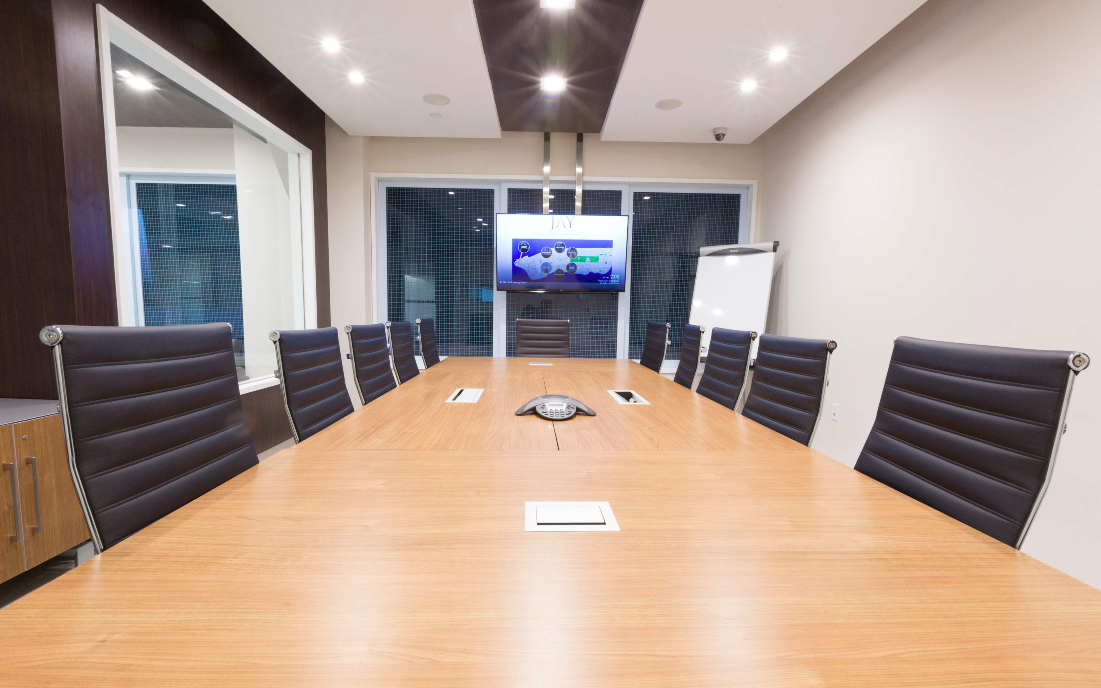 The image shows a modern conference room with a long wooden table, eight black leather chairs, a flat-screen TV mounted on the wall, and a whiteboard on a stand.