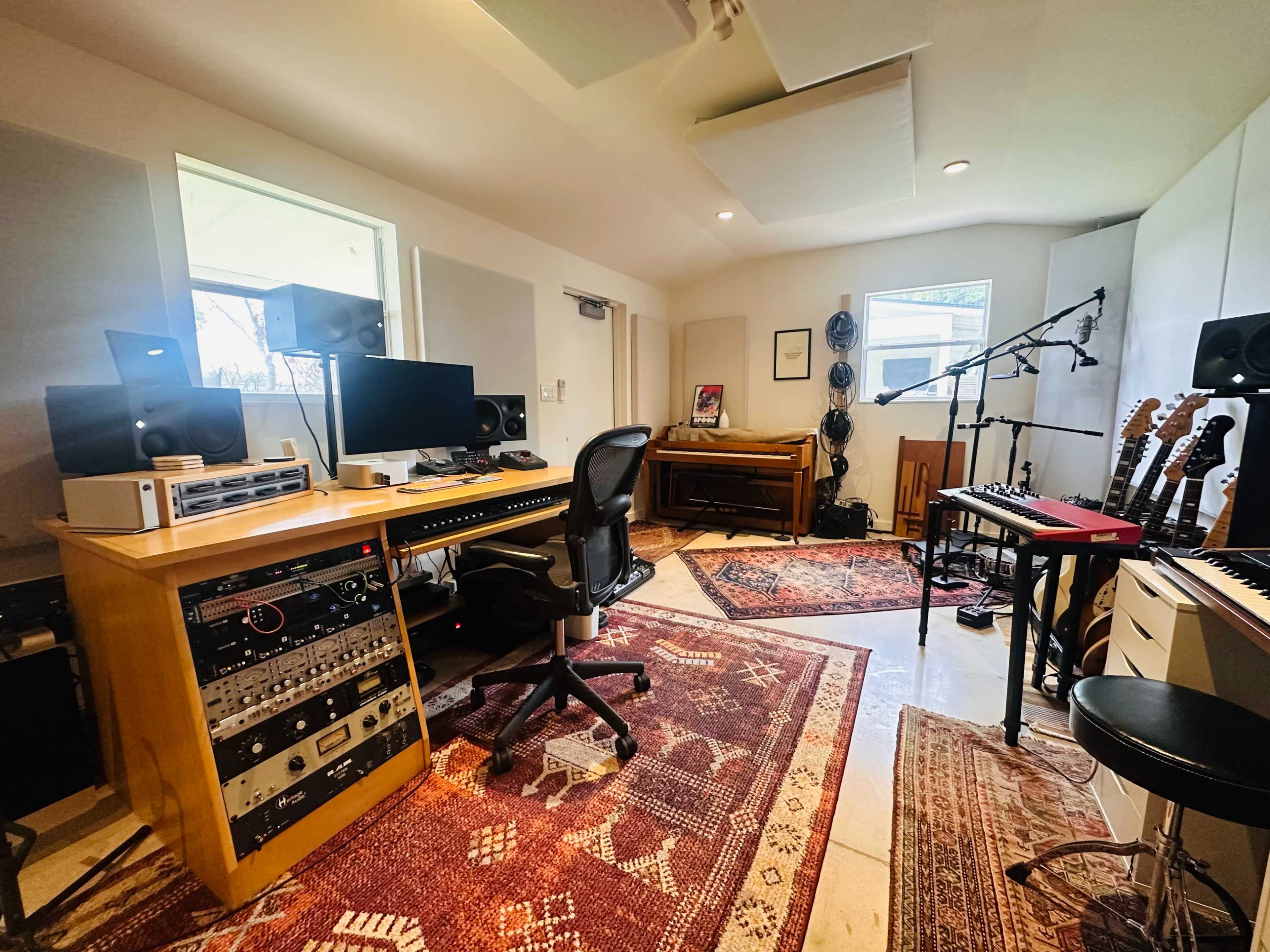 The image shows a recording studio featuring a desk with audio equipment, computer monitors, musical instruments, and rugs on the floor.