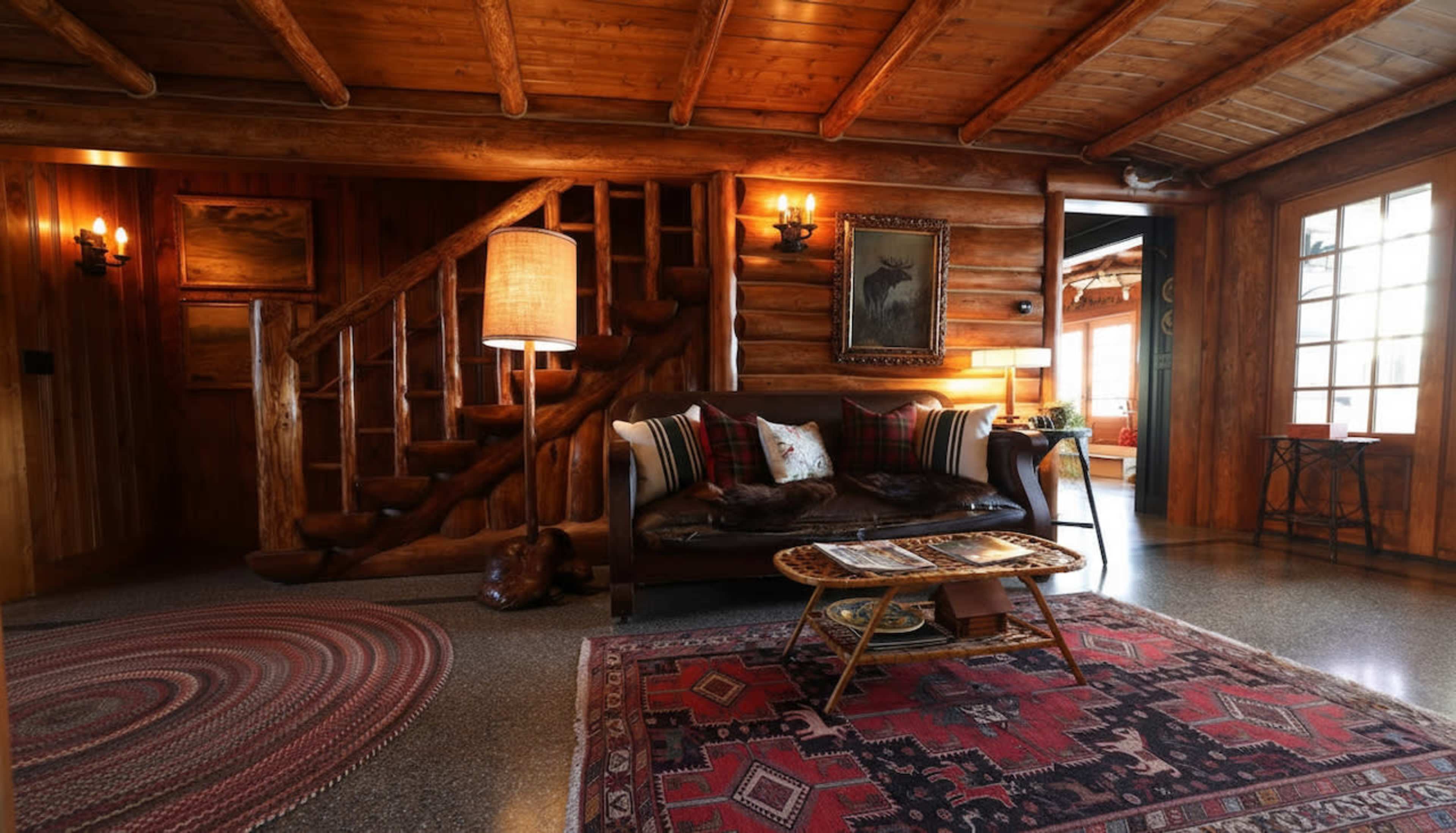 The image shows a rustic log cabin interior with a leather sofa, a decorative rug, a wooden staircase, and warm lighting.