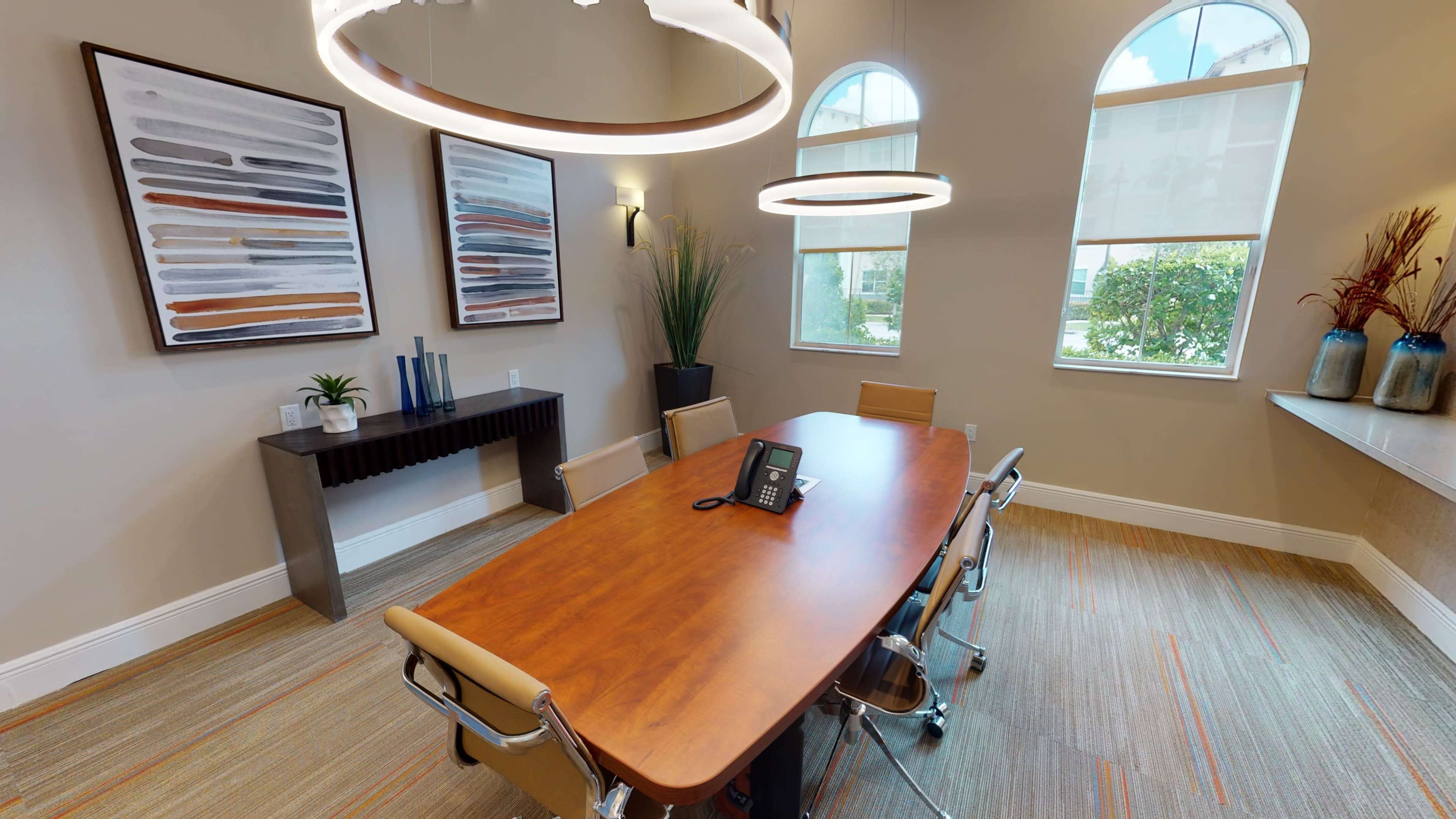 A modern conference room featuring a wooden table, four chairs, and abstract wall art, with large windows allowing natural light.