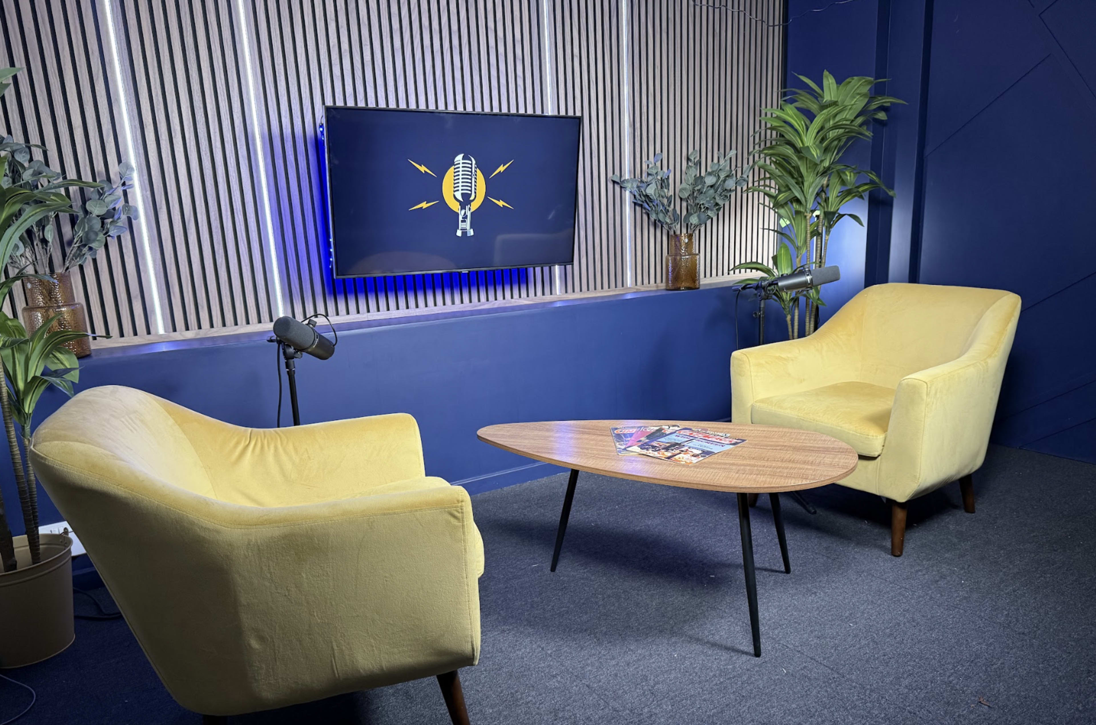 The image shows a modern interview set with two yellow armchairs, a wooden coffee table, and a wall-mounted television displaying a microphone logo.