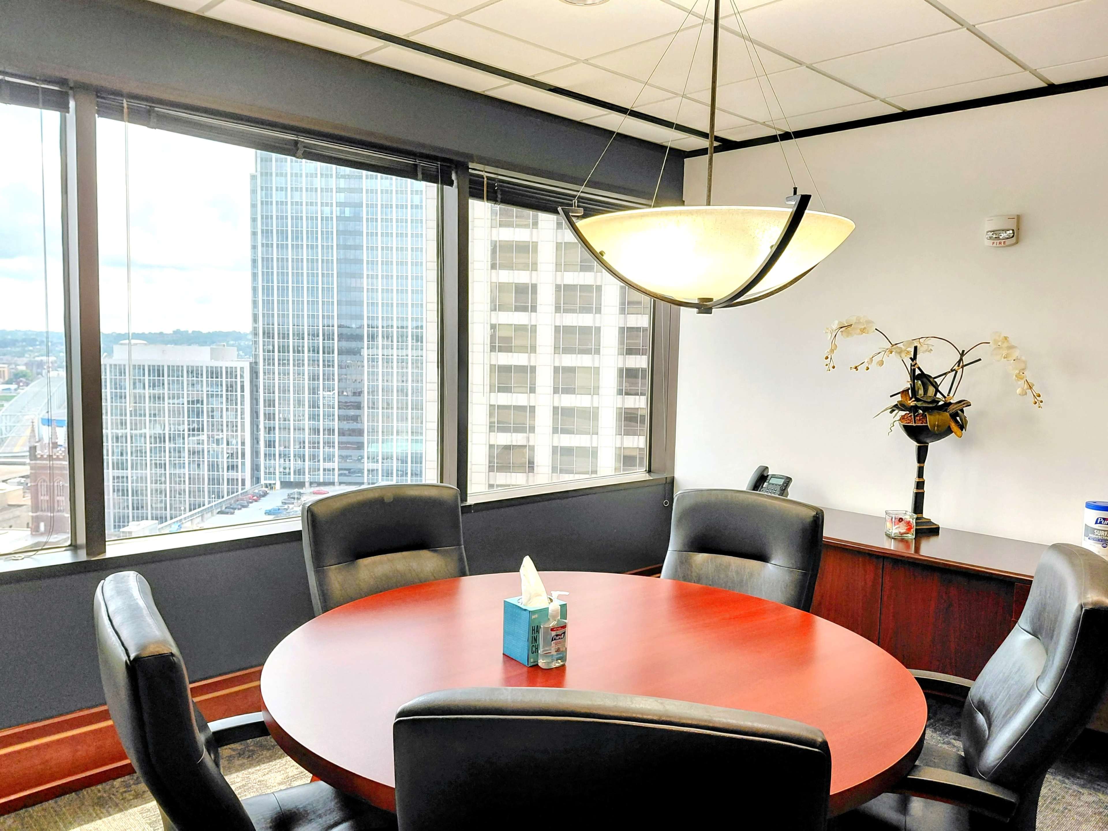 The image shows a modern office conference room with a round wooden table, black leather chairs, and large windows offering a view of a city skyline.
