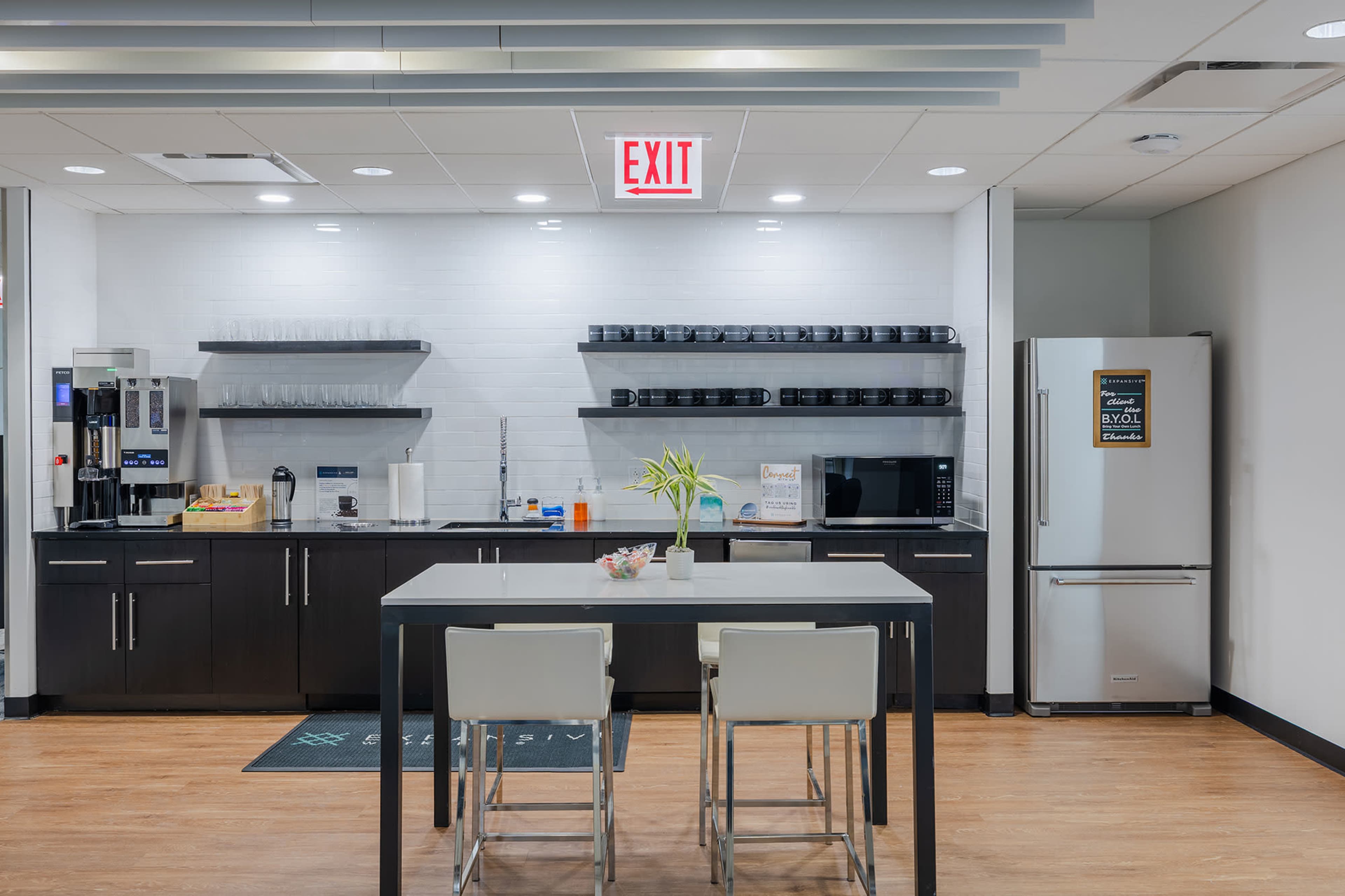 The image shows a modern kitchen area with a table and chairs, featuring shelves with mugs, a coffee machine, a microwave, and a refrigerator.